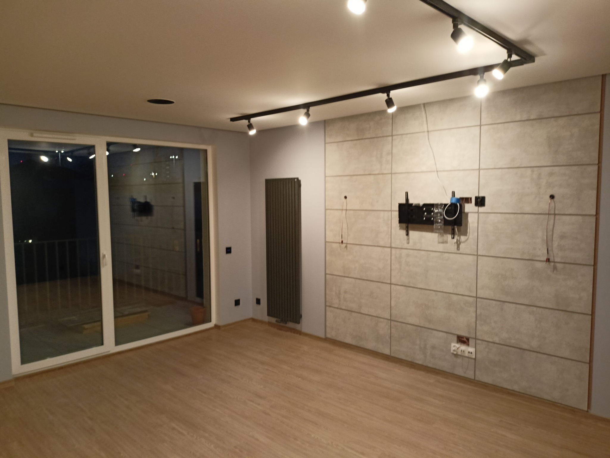 Nowoczesne wnętrze po remoncie: parkiet, szara ściana z panelami, grzejnik i oświetlenie szynowe. Widok na okno balkonowe i elementy montażu TV.