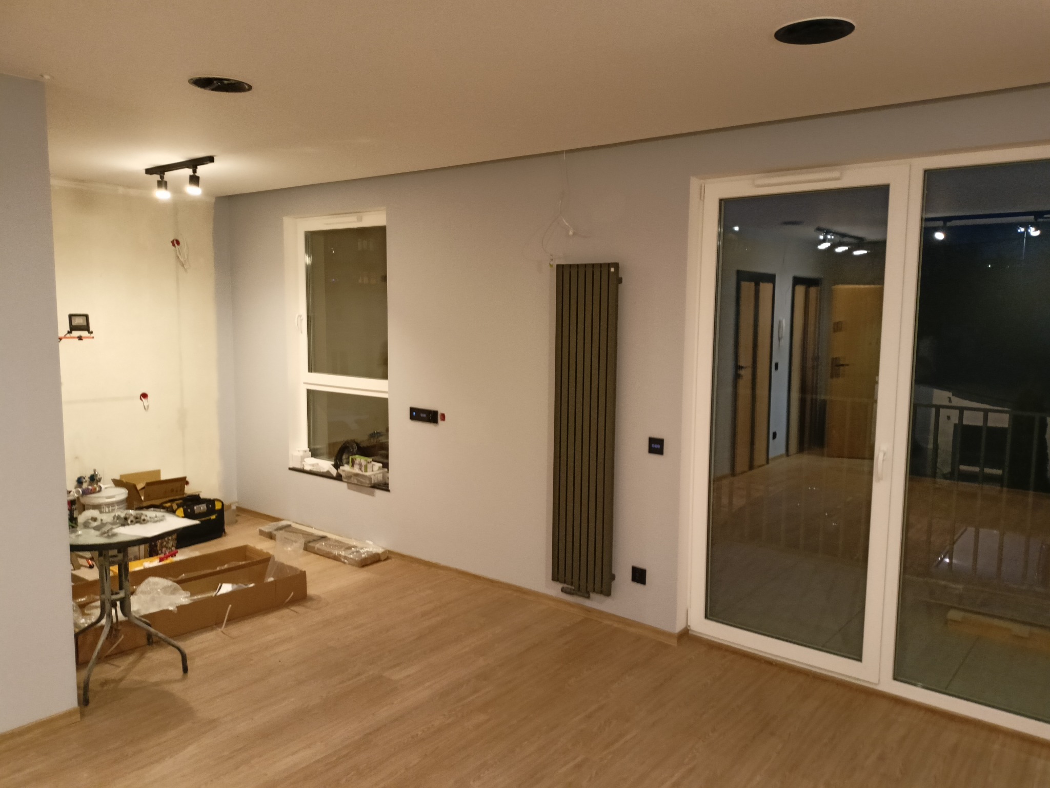 Wnętrze w trakcie remontu: widoczne narzędzia, kartony, grzejnik na ścianie, okno i drzwi balkonowe. Podłoga wyłożona panelami. Częściowo pomalowane ściany na szaro.