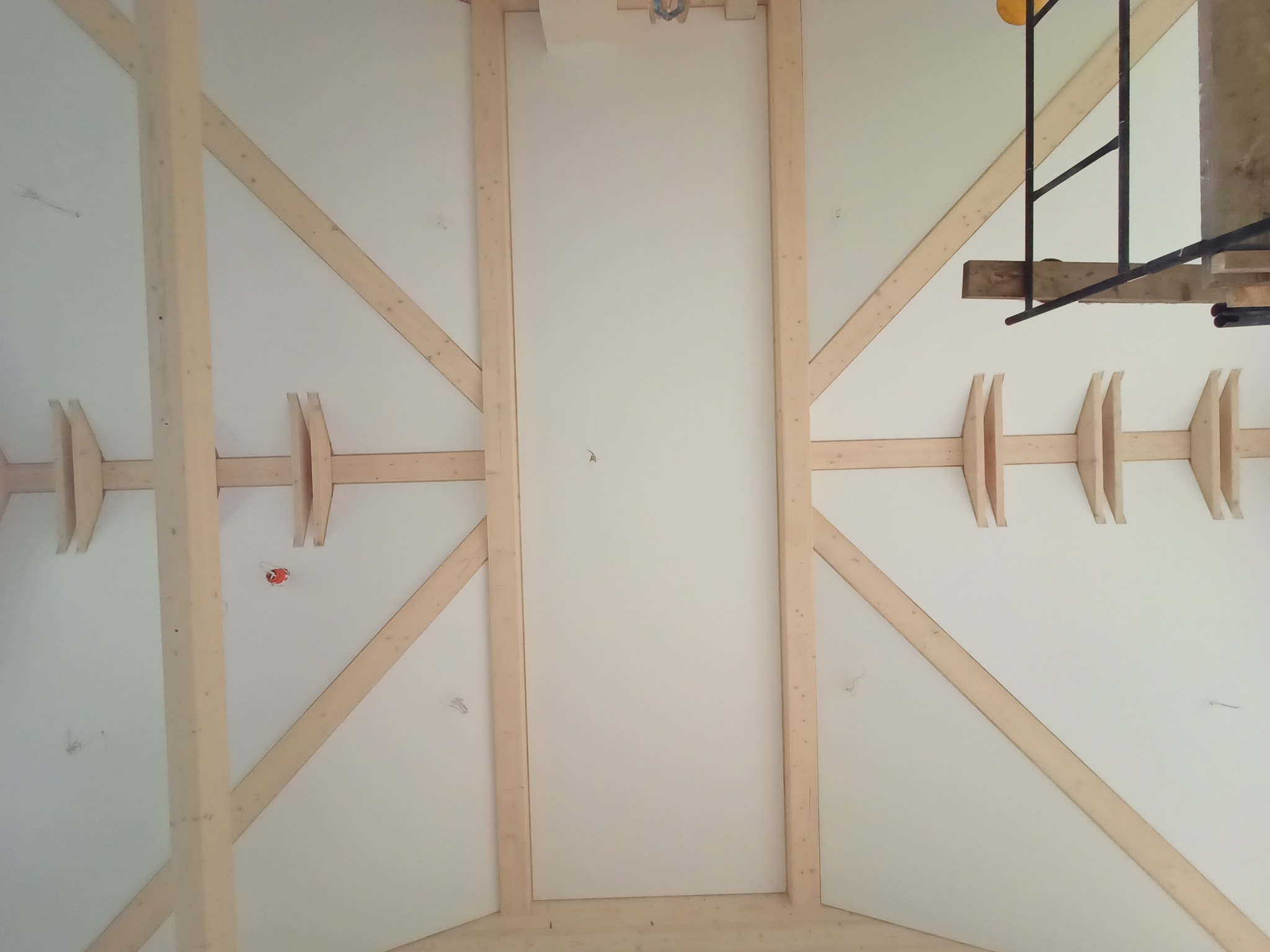 Drewniana konstrukcja stropu z widocznymi belkami i elementami mocującymi, białe wypełnienie między belkami, fragment rusztowania w prawym górnym rogu.