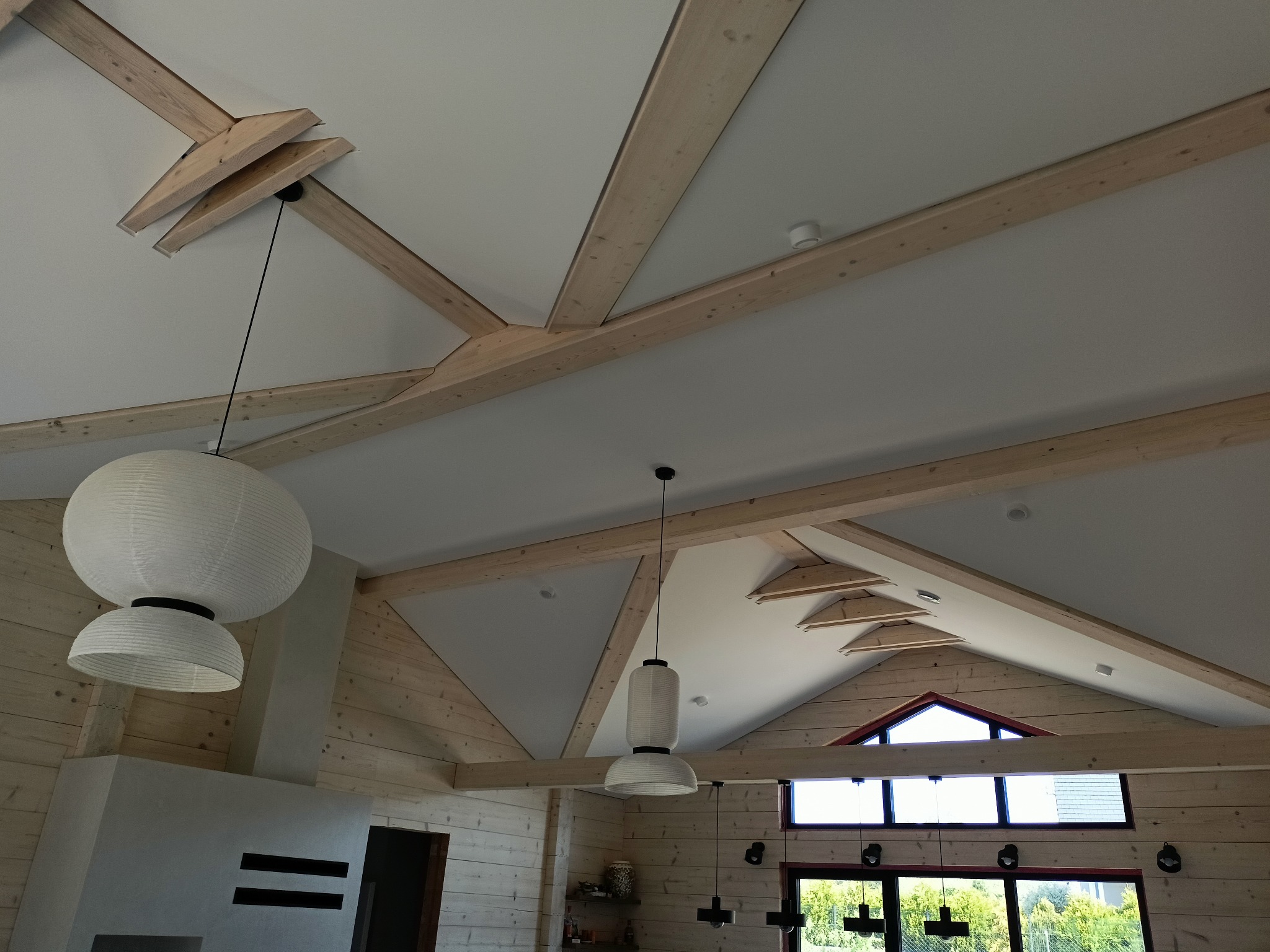 Drewniany sufit z widocznymi belkami i lampami w stylu japońskim. Jasne drewno, białe ściany i sufit. Widok na okno i zieleń na zewnątrz. Nowoczesny design.