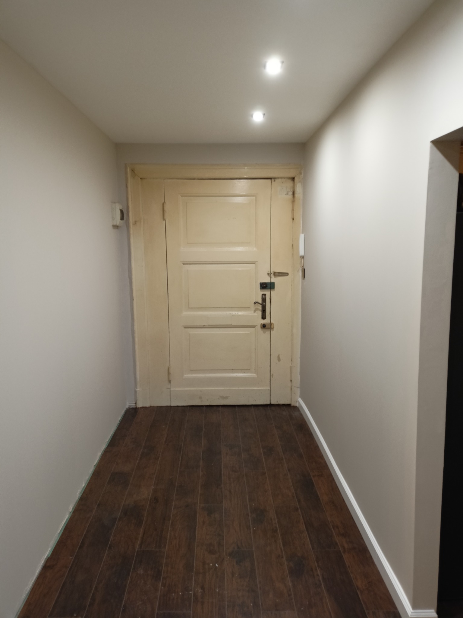 Korytarz po remoncie z nową podłogą imitującą drewno, świeżo pomalowanymi ścianami w odcieniach szarości i odnowionymi, kremowymi drzwiami z widocznymi śladami użytkowania.