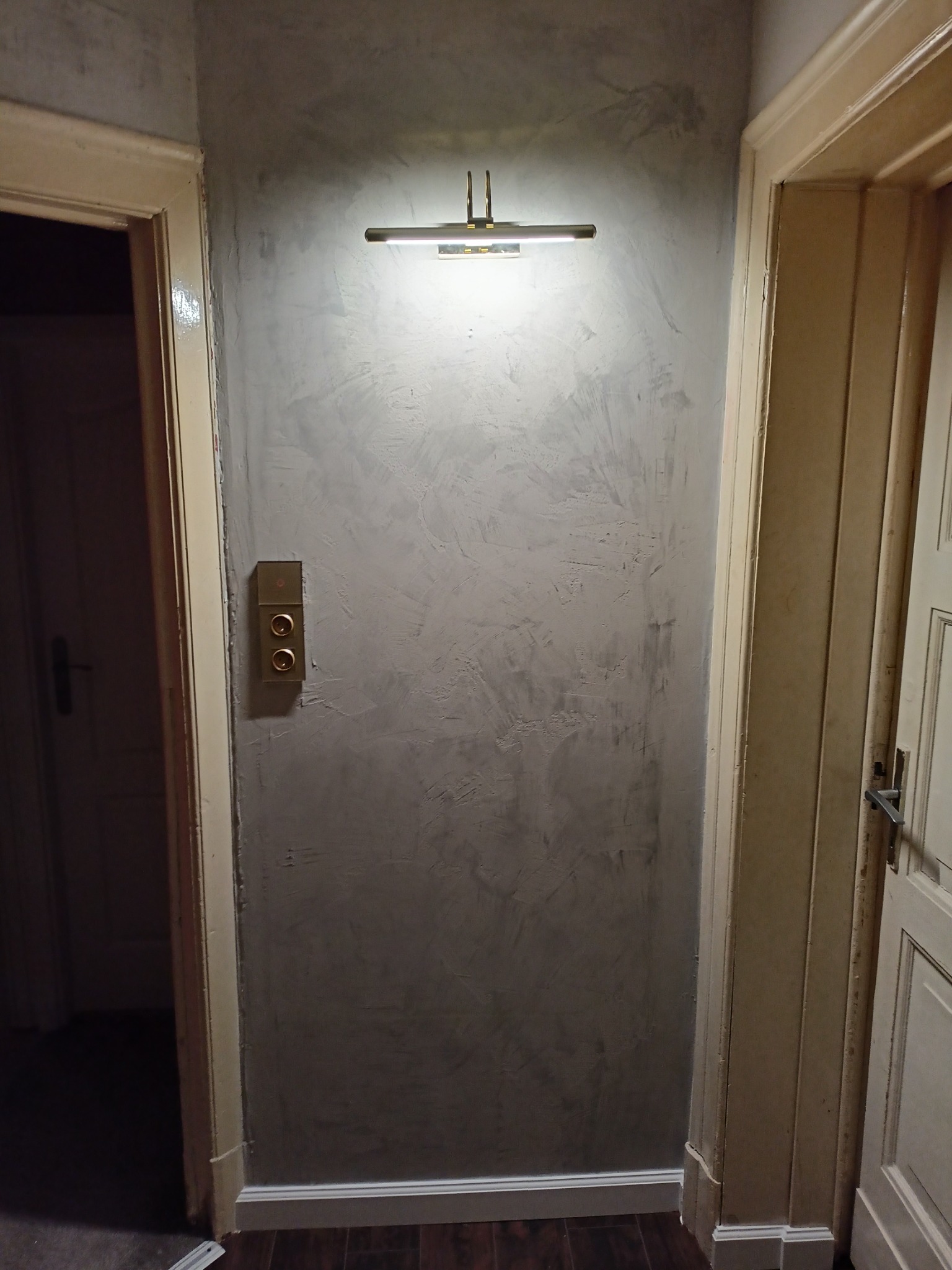 Ściana wykończona szarą, dekoracyjną fakturą, oświetlona podłużnym kinkietem LED, z widocznym włącznikiem światła i fragmentami dwóch ościeżnic drzwiowych.