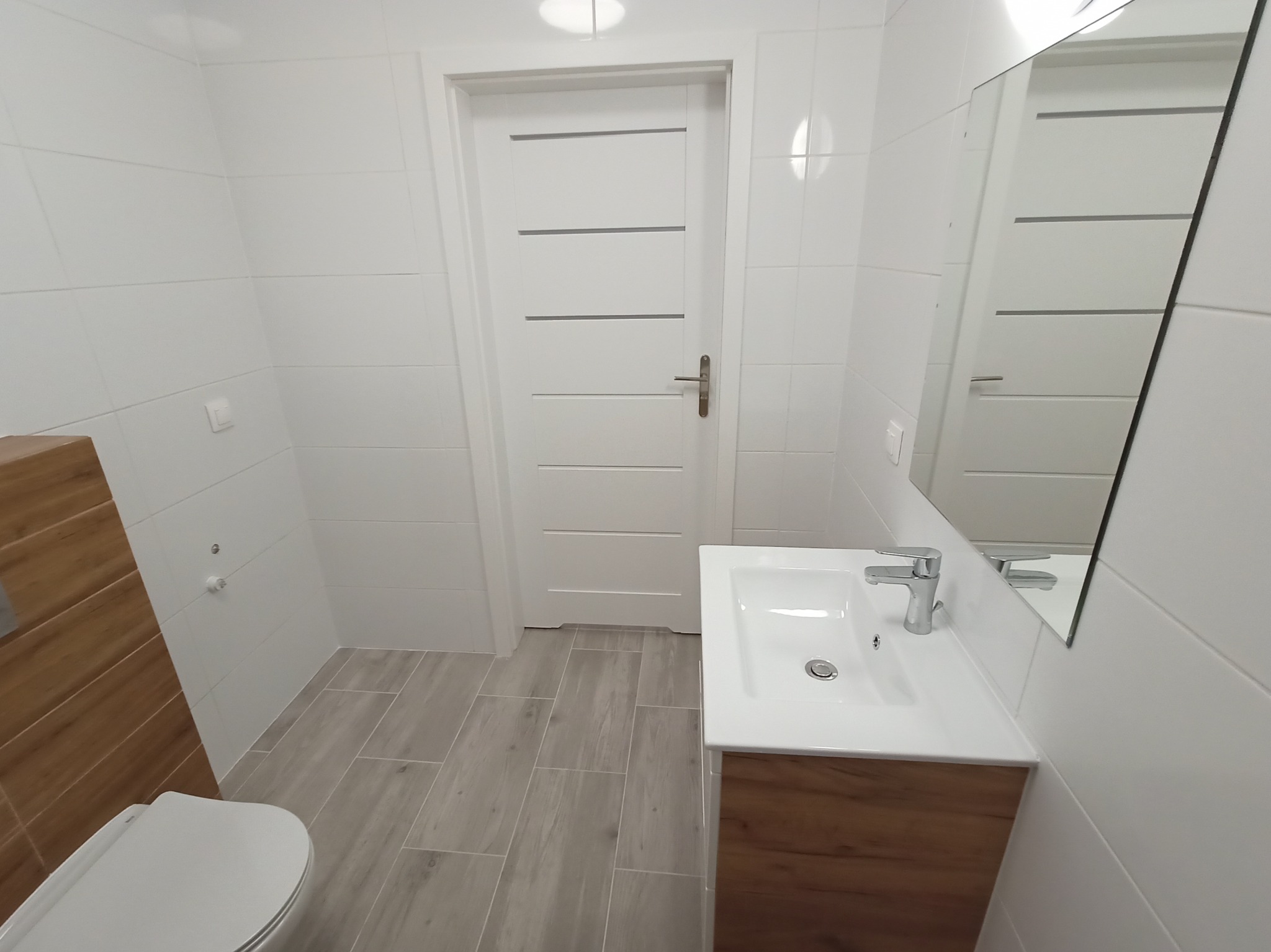 Nowoczesna łazienka z białymi płytkami, drewnopodobną szafką pod umywalką i szarą podłogą, widok na białe drzwi z poziomymi frezami.