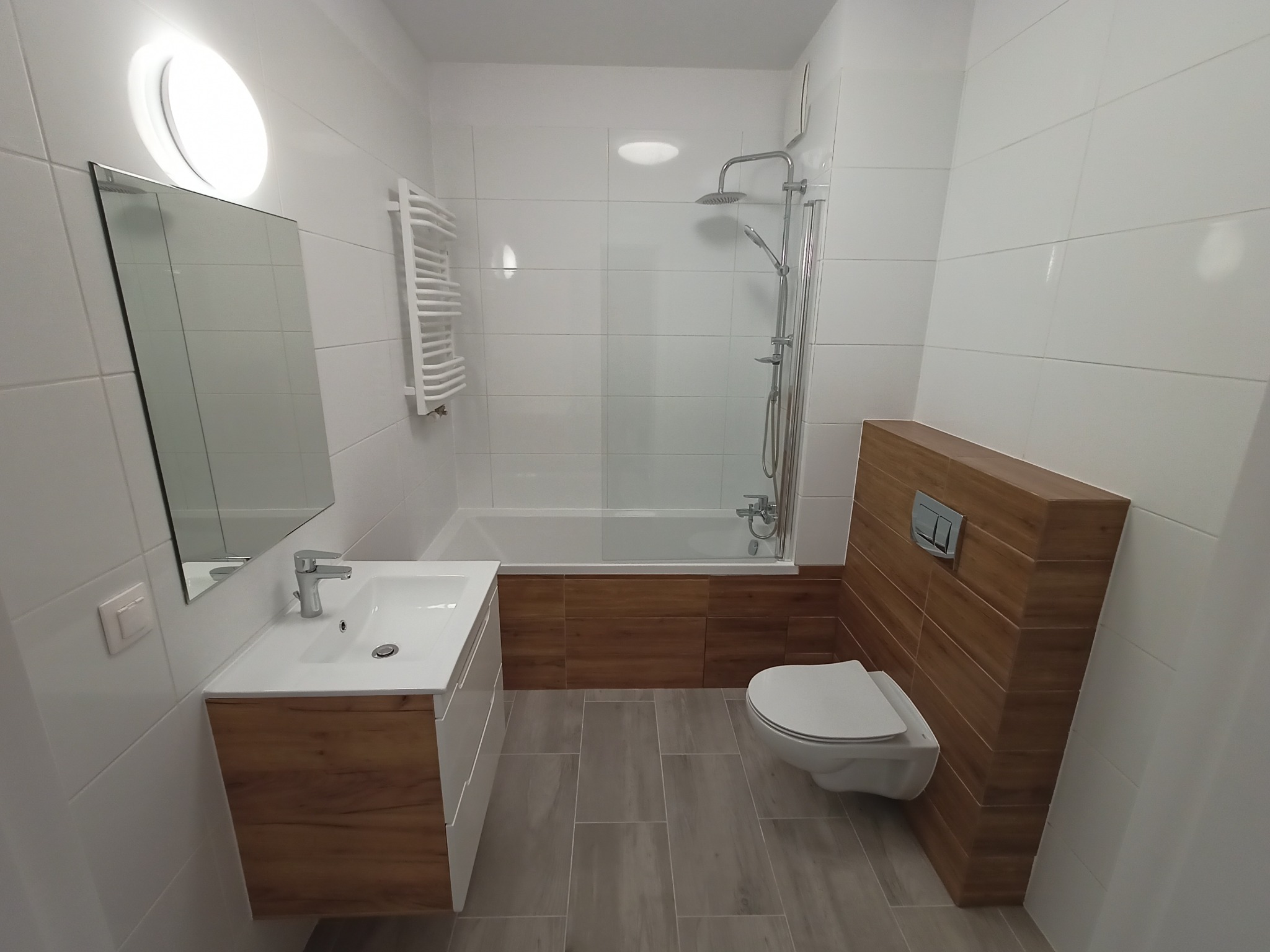 Nowoczesna łazienka z białymi płytkami, zabudowaną wanną z prysznicem, podwieszaną toaletą z obudową z paneli drewnopodobnych, szafką pod umywalkę z frontami w kolorze drewna i szarą podłogą...