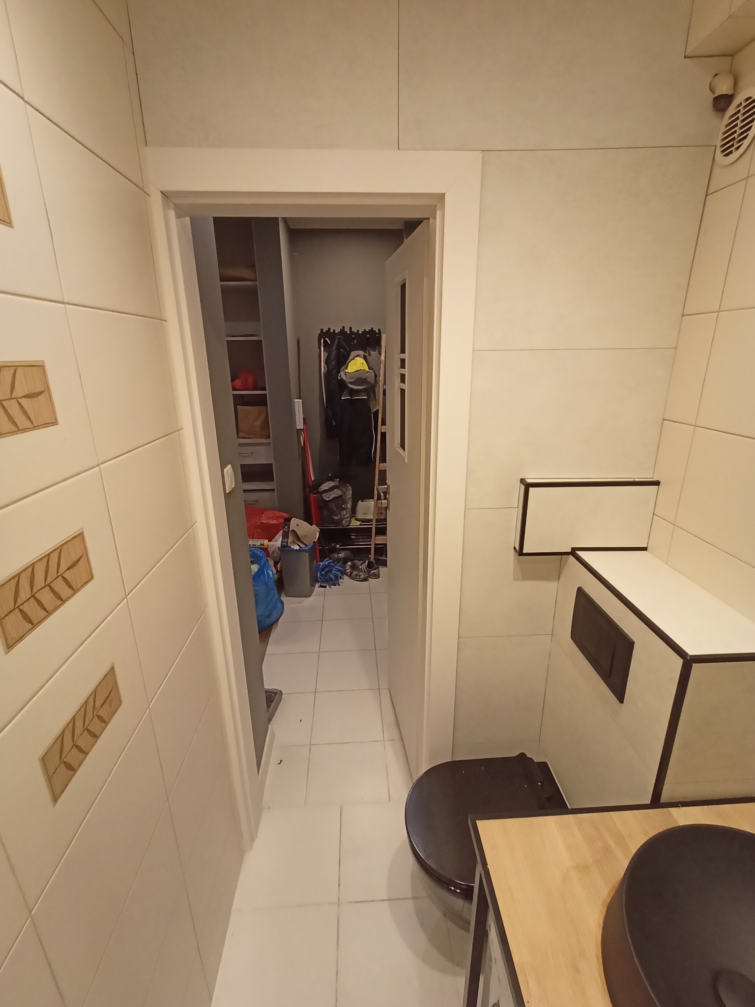 Wnętrze łazienki z beżowymi płytkami z drewnianymi wstawkami, widok na otwarte drzwi do pomieszczenia gospodarczego z regałem i rzeczami.