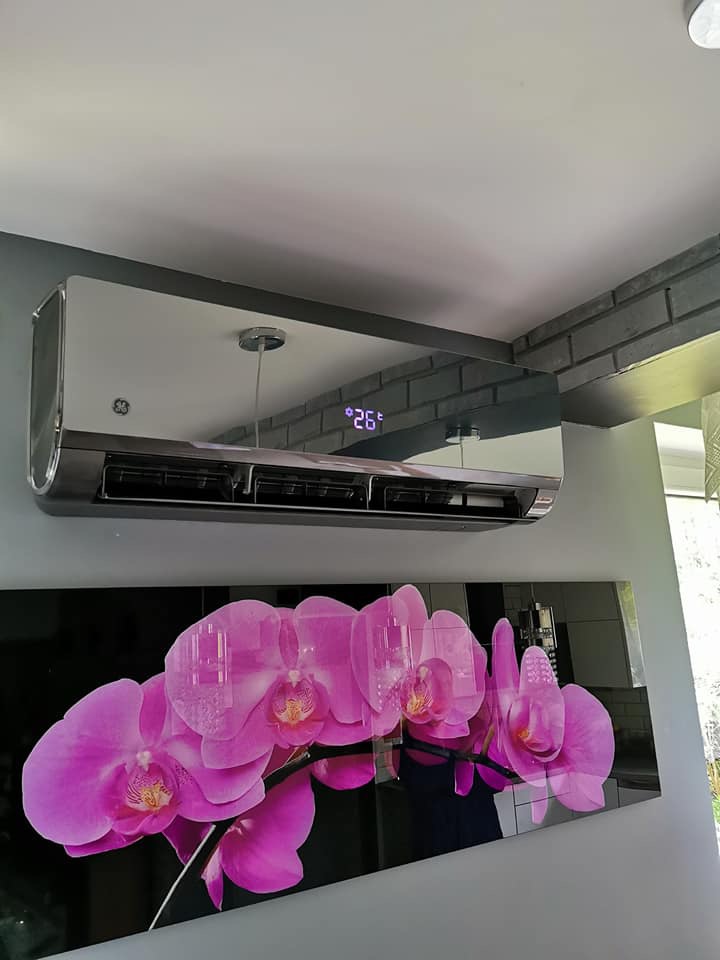Nowoczesny, chromowany klimatyzator ścienny marki GE z wyświetlaczem temperatury 26 stopni Celsjusza, zamontowany w pomieszczeniu z obrazem przedstawiającym różowe orchidee.