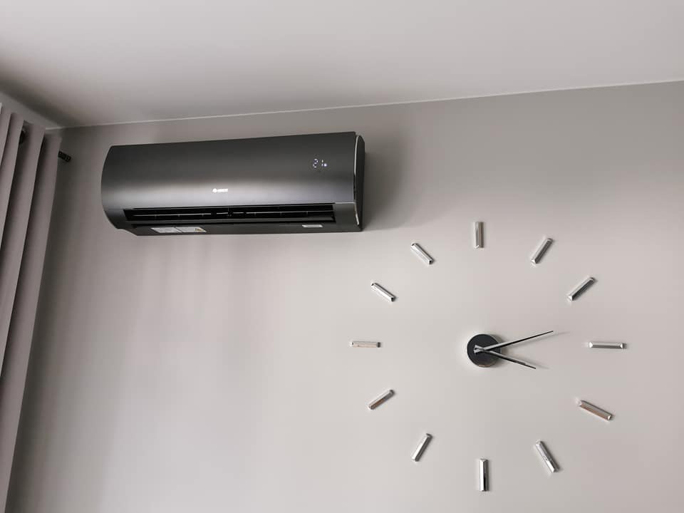 Czarny klimatyzator marki Gree zamontowany na jasnej ścianie w pokoju, obok dekoracyjny zegar ścienny.