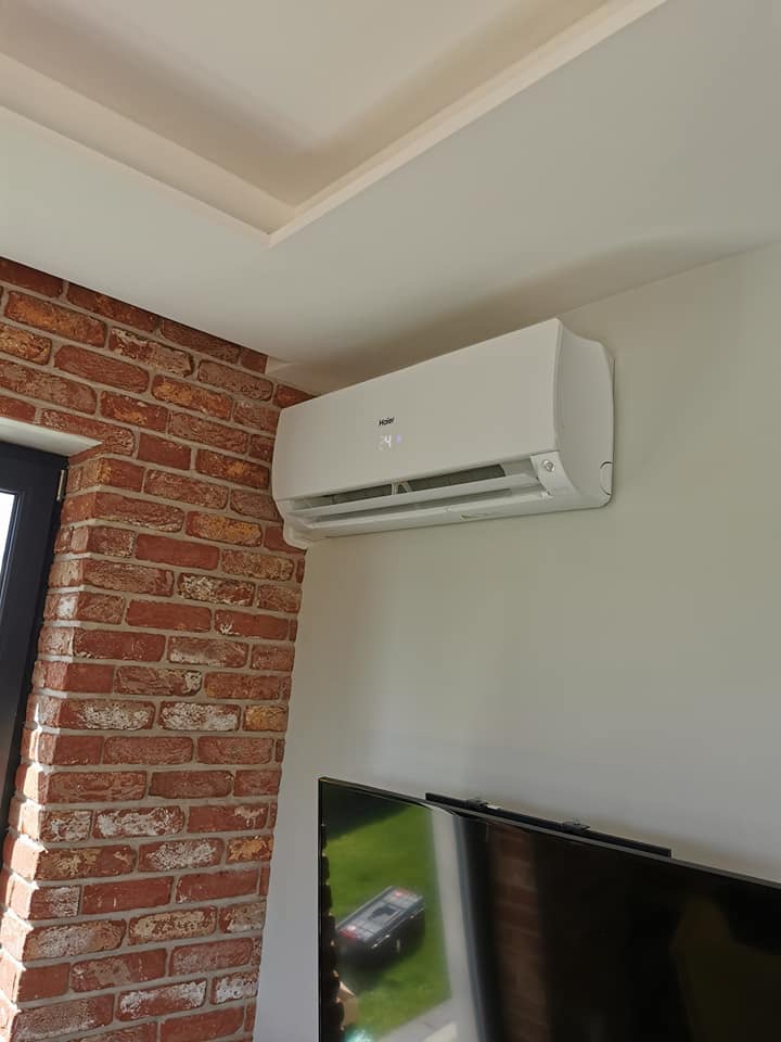 Biały klimatyzator Haier zamontowany na ścianie w pomieszczeniu z ceglaną kolumną i telewizorem.