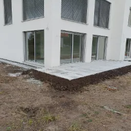 Nowoczesny dom z jasną elewacją i tarasem z szarych płyt, obrzeże tarasu obsypane ziemią przygotowane do nasadzeń, widoczne okna przesuwne z szarymi ramami i żaluzjami zewnętrznymi.