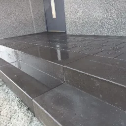 Nowoczesne, ciemnoszare schody wejściowe wyłożone kostką brukową, z widocznym podłożem z grysu i elewacją budynku w tle.