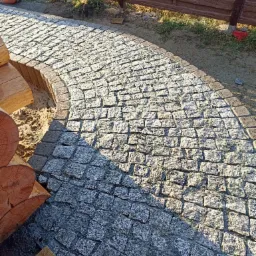 Ścieżka z szarej kostki granitowej, obrzeża z ciemniejszych kostek, widoczne elementy drewniane i fragment brązowego ogrodzenia.