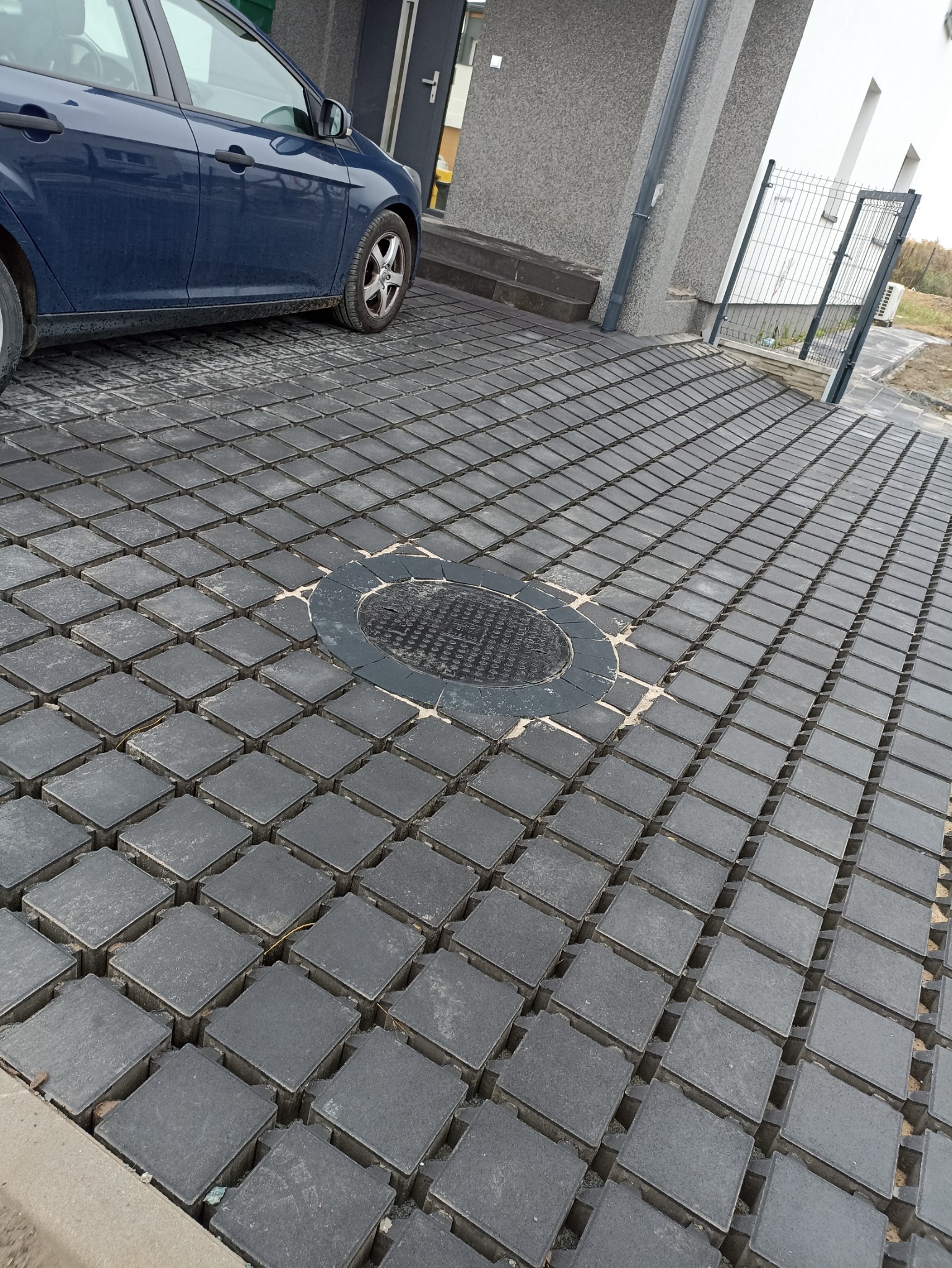 Szara, przepuszczalna nawierzchnia z kostki brukowej z widoczną studzienką kanalizacyjną i zaparkowanym samochodem osobowym.