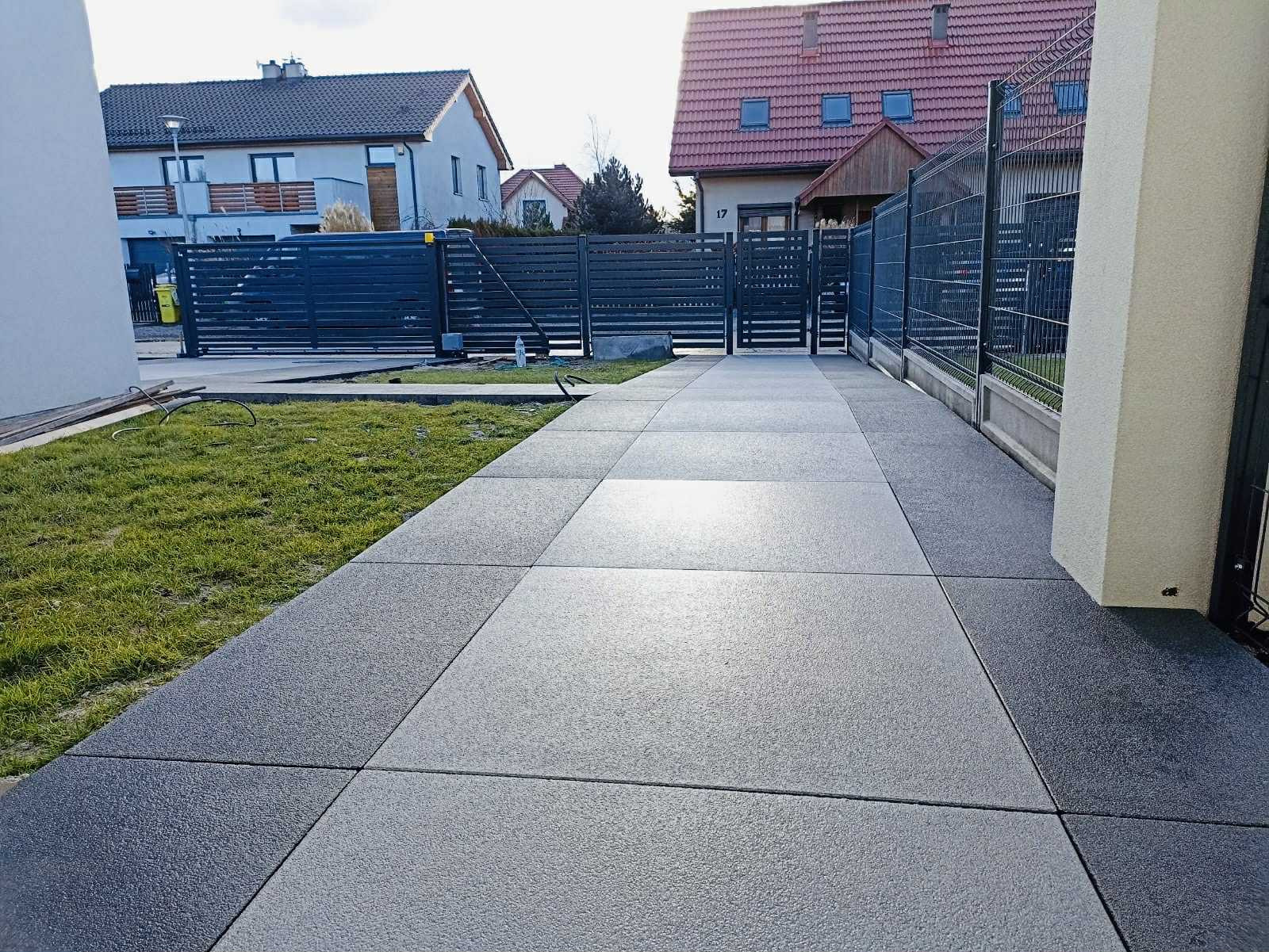 Nowoczesny podjazd z szarej kostki brukowej o gładkiej fakturze, ułożonej w geometryczny wzór. Widoczny fragment trawnika i ogrodzenie posesji.