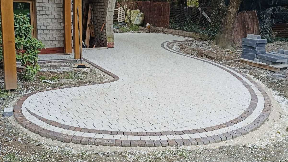 Kremowa kostka brukowa ułożona w łukowaty wzór z obramowaniem z ciemniejszej kostki, tworząca ścieżkę przed budynkiem. Widoczne zapasy kostki brukowej.