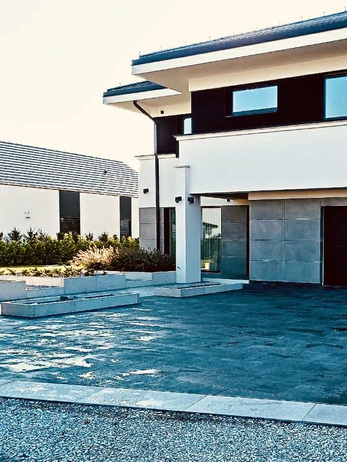 Nowoczesny podjazd z betonu architektonicznego i żwiru, minimalistyczne donice, dom w stylu modernistycznym z płaskim dachem w tle, jasne oświetlenie.