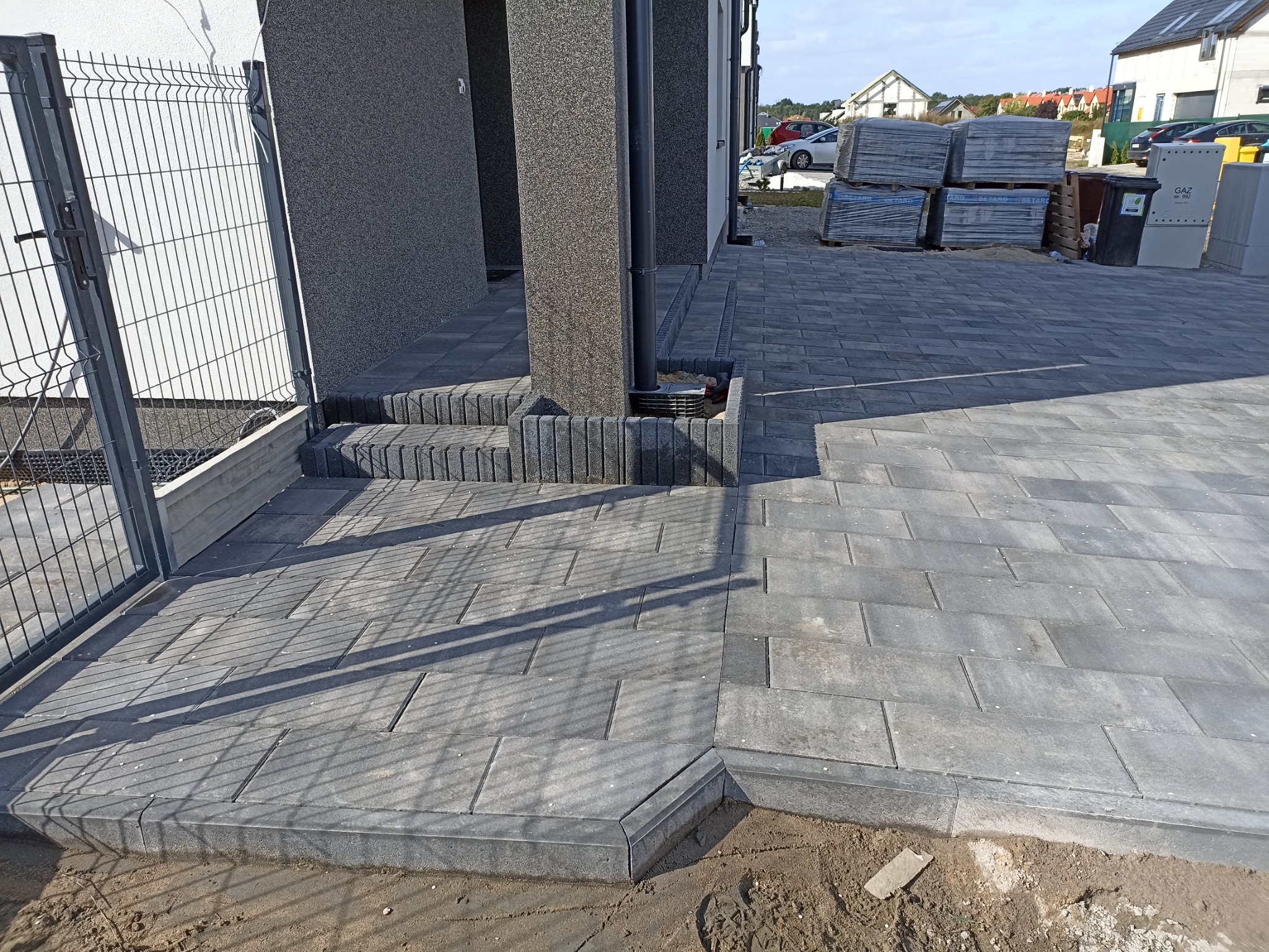 Nowo ułożona szara kostka brukowa prowadząca do wejścia domu z widocznymi schodami, obrzeżami i paletami kostki w tle.