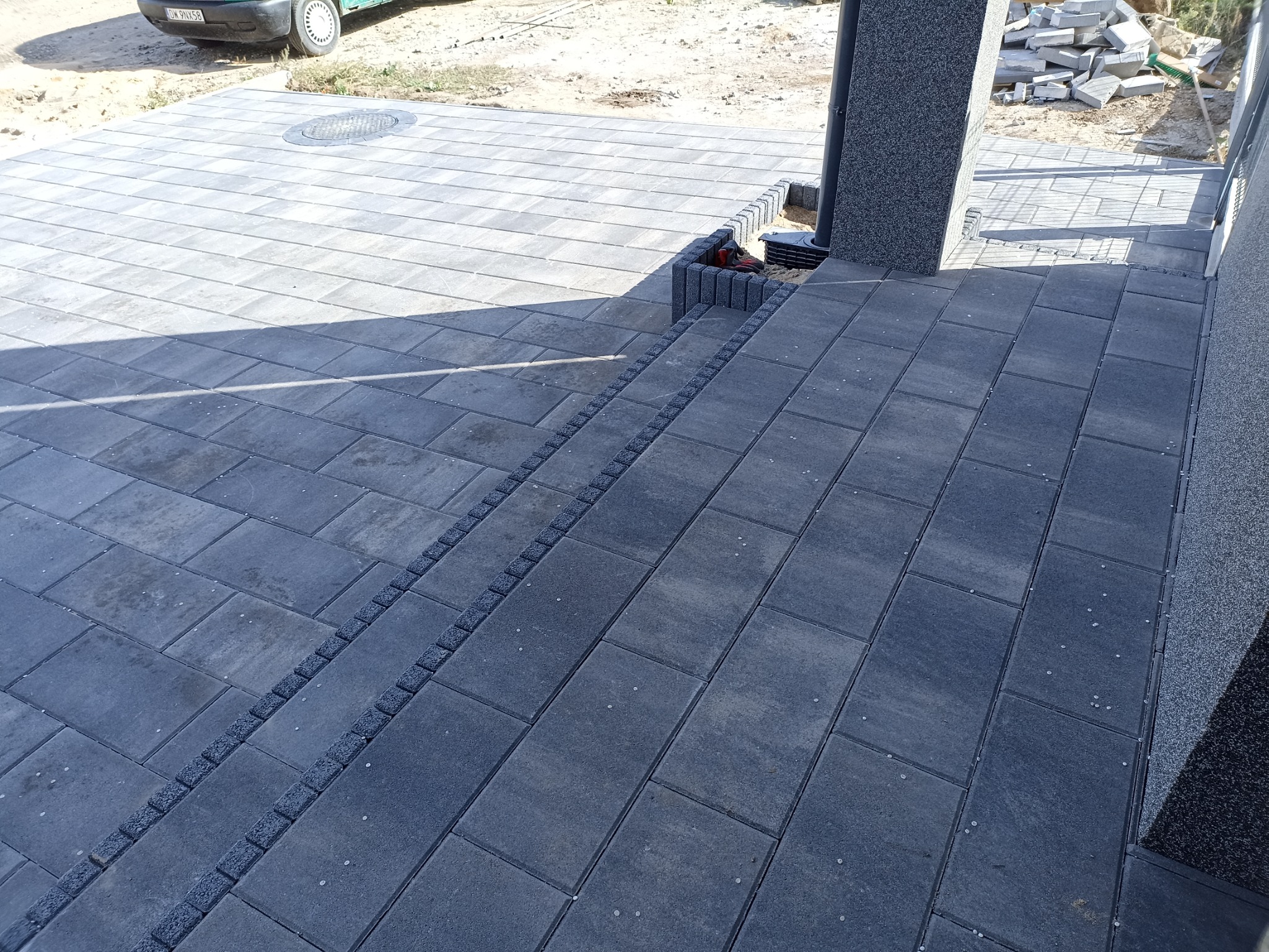 Ułożona, szara kostka brukowa tworząca schody i chodnik, z widocznym odwodnieniem liniowym i elementami wykończeniowymi z drobnej kostki, w tle zaparkowany samochód i sterta kostki brukowej.