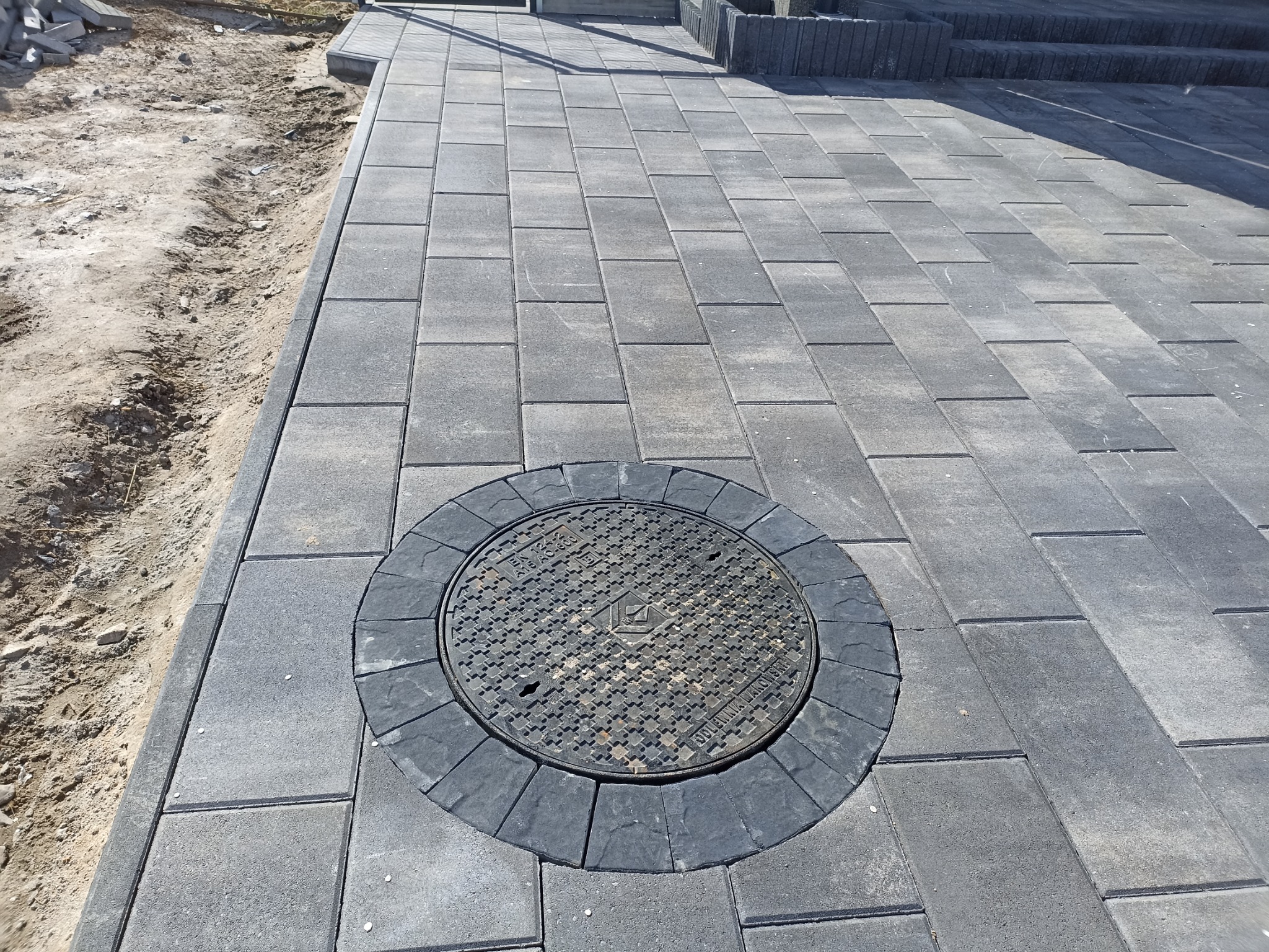 Nowa, szara kostka brukowa ułożona wokół studzienki kanalizacyjnej z ciemnoszarą obwódką, widoczny fragment niezakończonych prac ziemnych po lewej stronie.