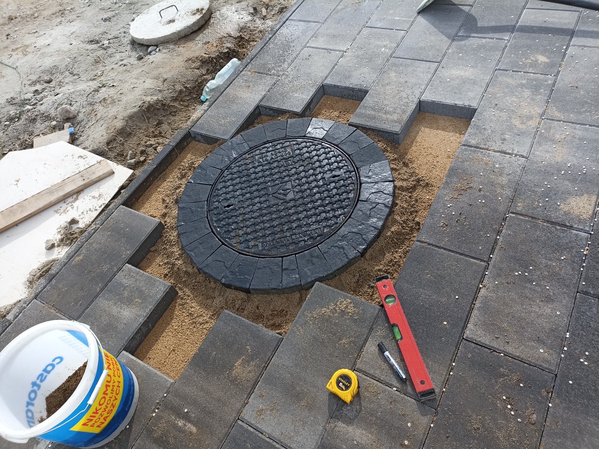Układanie ciemnoszarej kostki brukowej wokół okrągłej studzienki kanalizacyjnej z użyciem poziomicy, miarki i piasku.