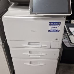 JANAS Kamil Janas - Biała kserokopiarka Ricoh z naklejką informacyjną o eksploatacji, serwisie i dostawie. Widoczne szuflady na papier i panel sterowania.
