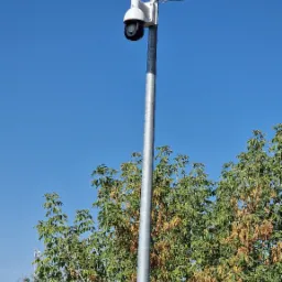 Kamera przemysłowa na słupie w Milejowicach, skierowana w górę. Widoczne okablowanie i drzewa w tle. Słoneczny dzień, błękitne niebo.