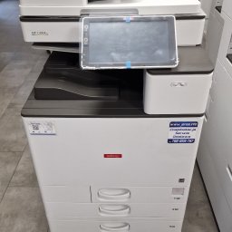 JANAS Kamil Janas - Biała kserokopiarka Nashuatec z czterema szufladami i panelem dotykowym, widoczna naklejka z danymi kontaktowymi serwisu. W tle inne urządzenia.