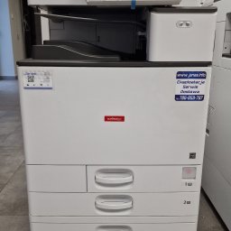 JANAS Kamil Janas - Wielofunkcyjna kserokopiarka Nashuatec MP C3004ex z naklejkami informacyjnymi firmy Janas, widok z przodu na szarej podłodze.