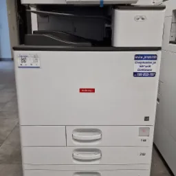 Wielofunkcyjna kserokopiarka Nashuatec MP C3004ex z naklejkami informacyjnymi firmy Janas, widok z przodu na szarej podłodze.