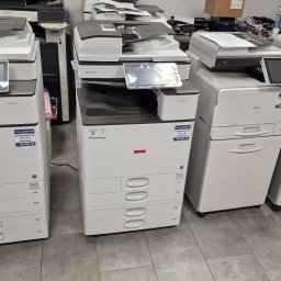 JANAS Kamil Janas - Rząd używanych, wielofunkcyjnych kserokopiarek A4, różnych modeli i marek, z widocznymi panelami dotykowymi, w pomieszczeniu biurowym przed serwisem.