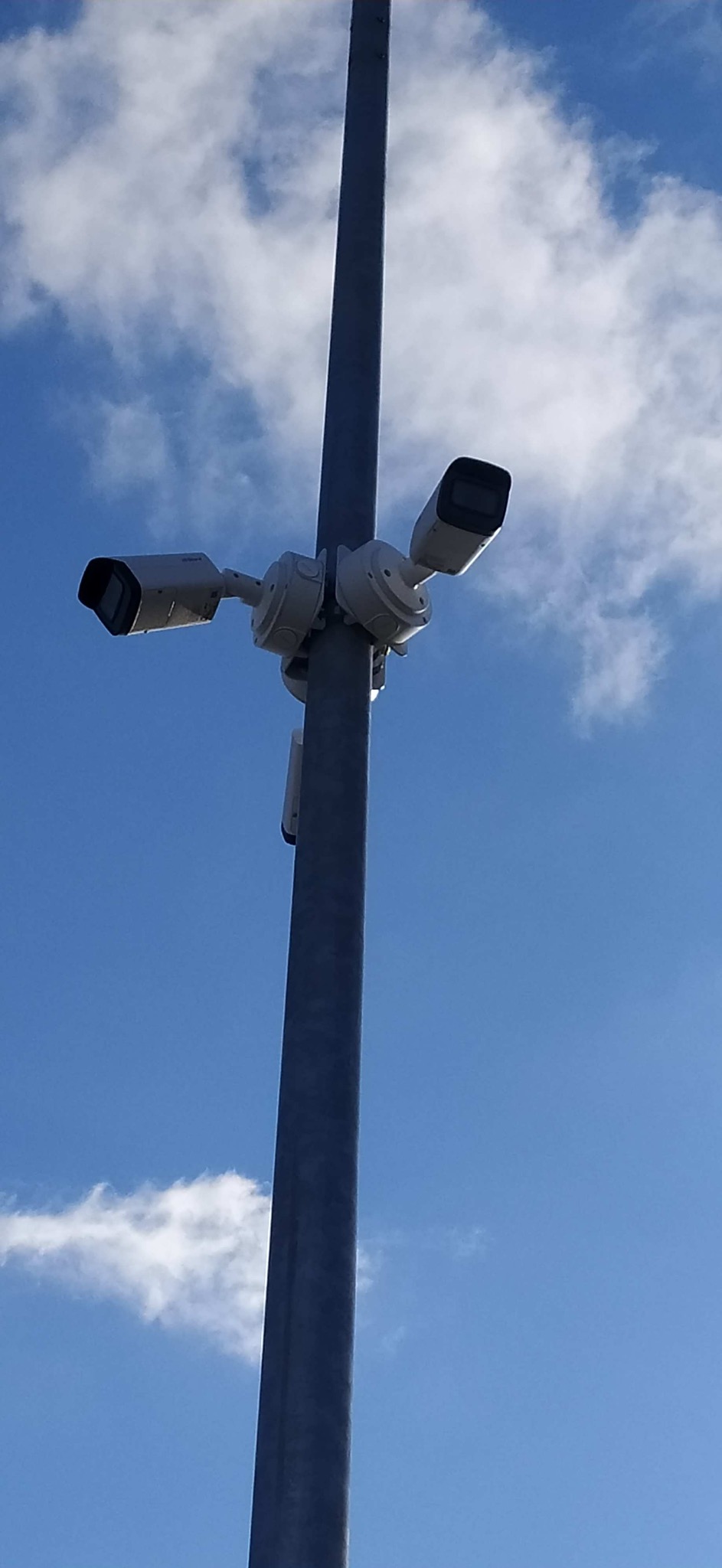 Słup z trzema kamerami monitoringu skierowanymi w różne strony na tle błękitnego nieba z chmurami. Widok z dołu na zainstalowany system.