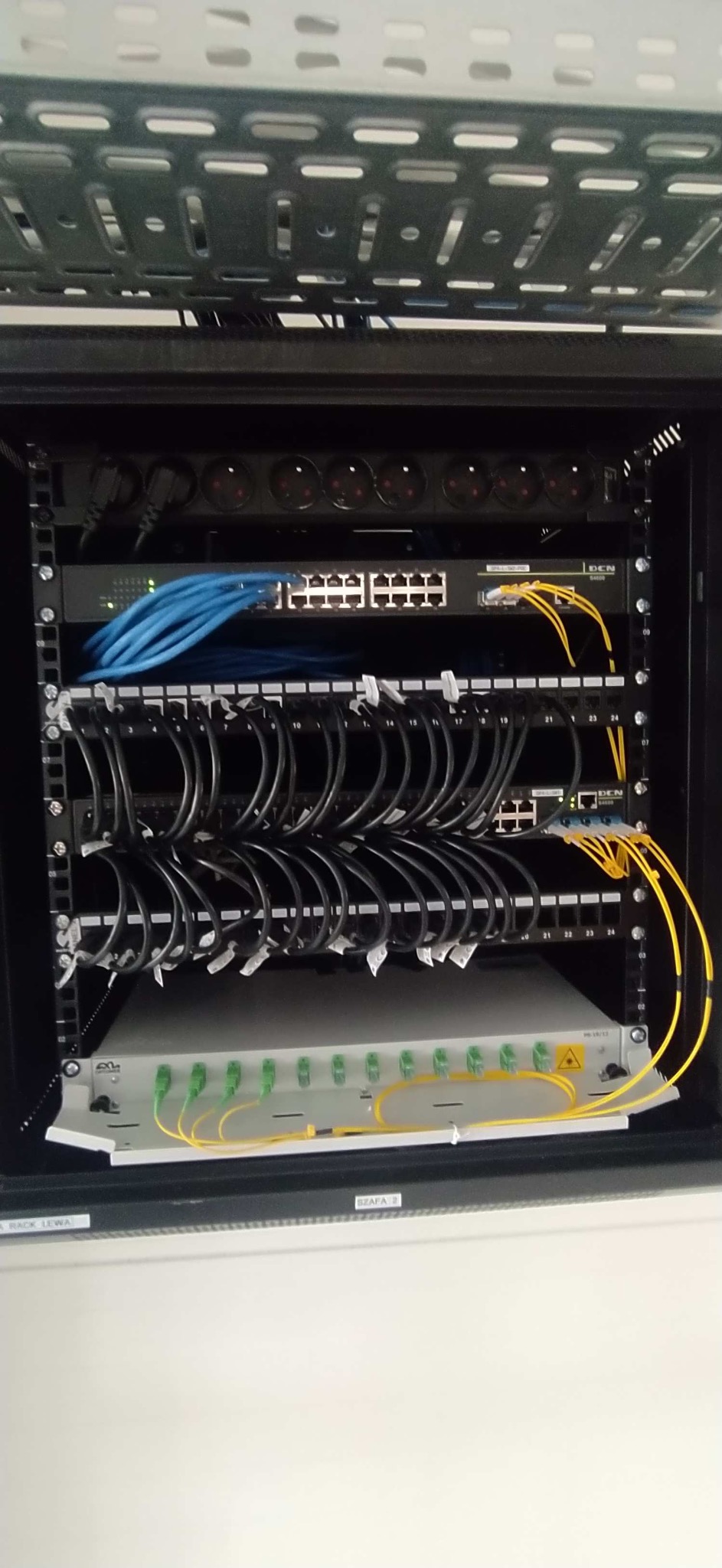 Zamknięta szafa rack z uporządkowanymi kablami sieciowymi (patchcordy) i urządzeniami, w tym switch, listwa zasilająca i moduł światłowodowy. Kable czarne z białymi etykietami.