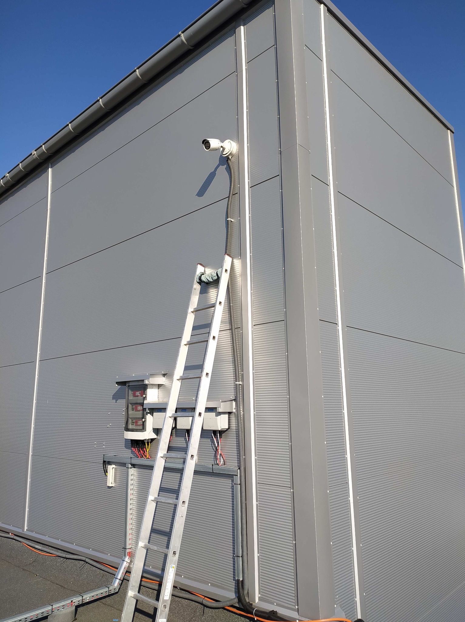 Montaż kamery monitoringu na szarej elewacji budynku. Aluminiowa drabina oparta o ścianę, widoczne kable i skrzynka elektryczna. Błękitne niebo w tle.