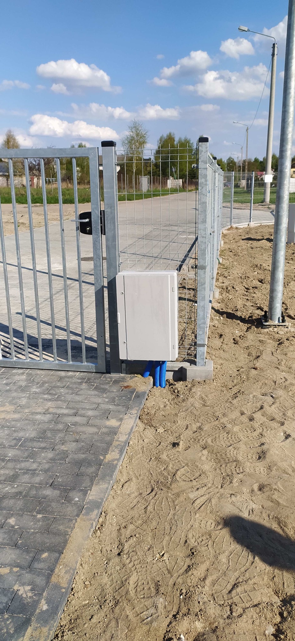 Szara skrzynka sterownicza zamontowana na metalowym ogrodzeniu panelowym z niebieskimi rurami osłonowymi kabli, widok z boku na kostkę brukową i piaszczyste podłoże.
