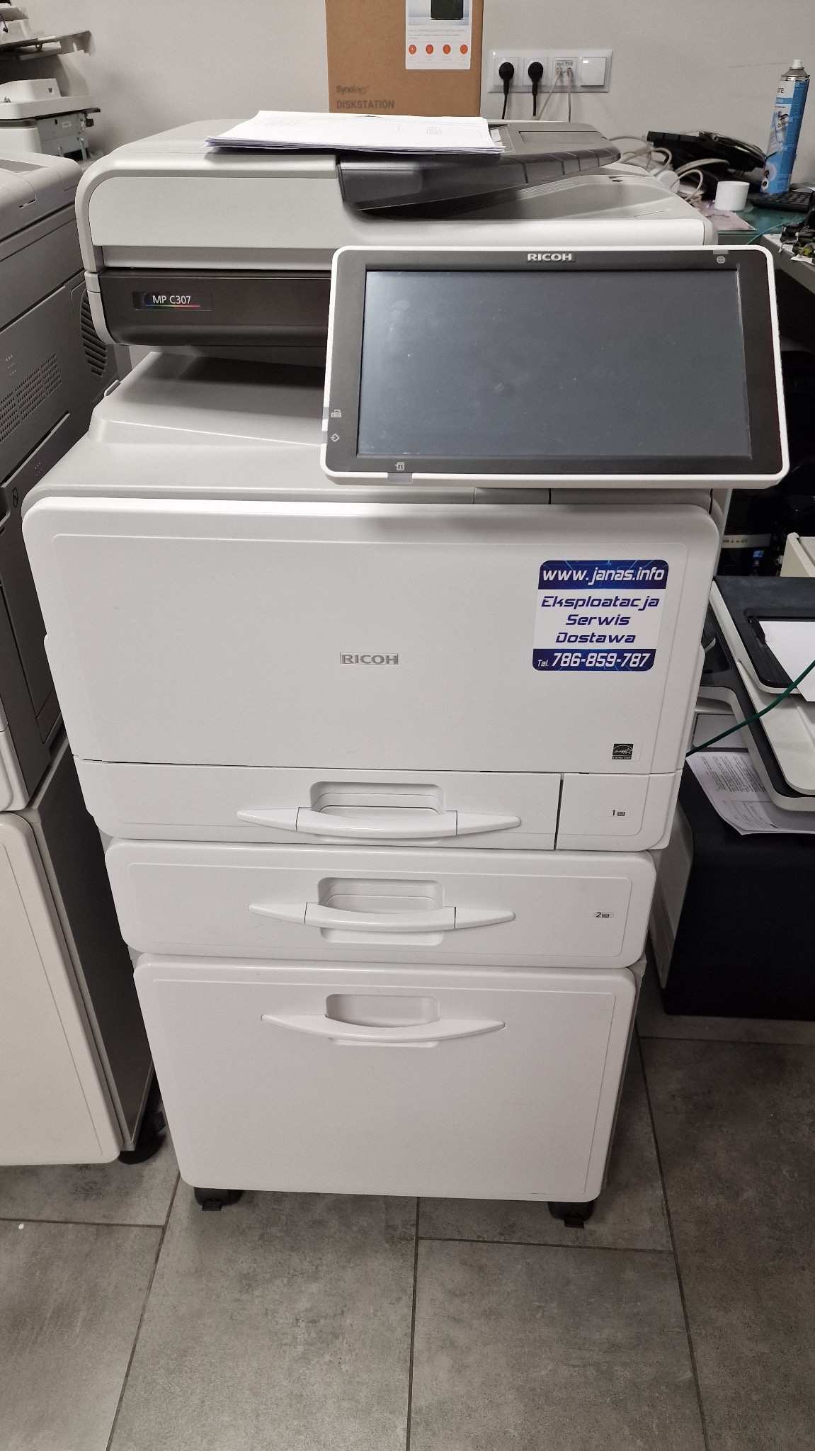 Biała kserokopiarka Ricoh z naklejką informacyjną o eksploatacji, serwisie i dostawie. Widoczne szuflady na papier i panel sterowania.