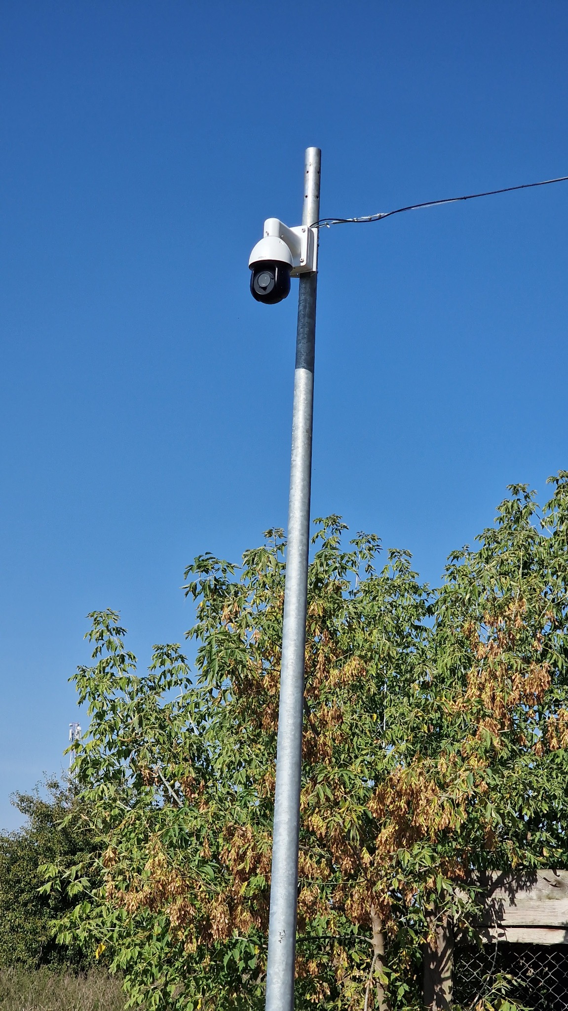 Kamera przemysłowa na słupie w Milejowicach, skierowana w górę. Widoczne okablowanie i drzewa w tle. Słoneczny dzień, błękitne niebo.