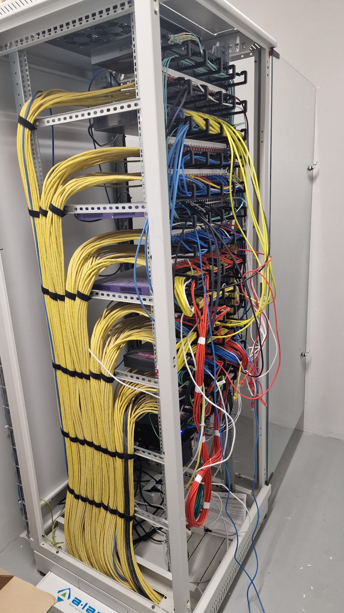 Szafa rack z uporządkowanymi kablami ethernetowymi w kolorach żółtym, niebieskim i czerwonym. Kable po lewej stronie spięte czarnymi opaskami.