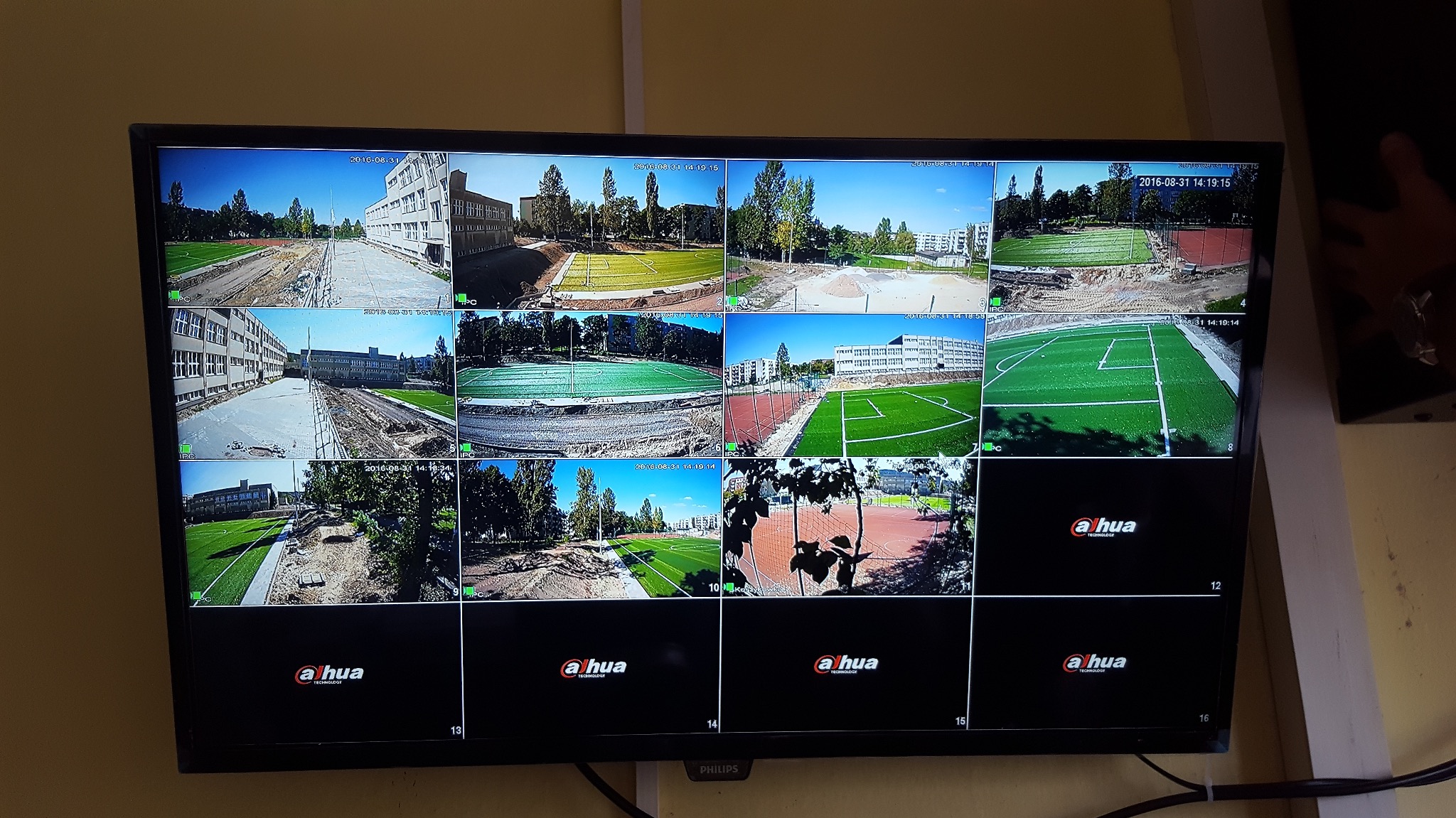 Ekran monitoringu Dahua pokazuje szesnaście kamer z widokiem na budowę boiska szkolnego, z datami w rogu. Widoczne budynki szkolne i postęp prac nad murawą.