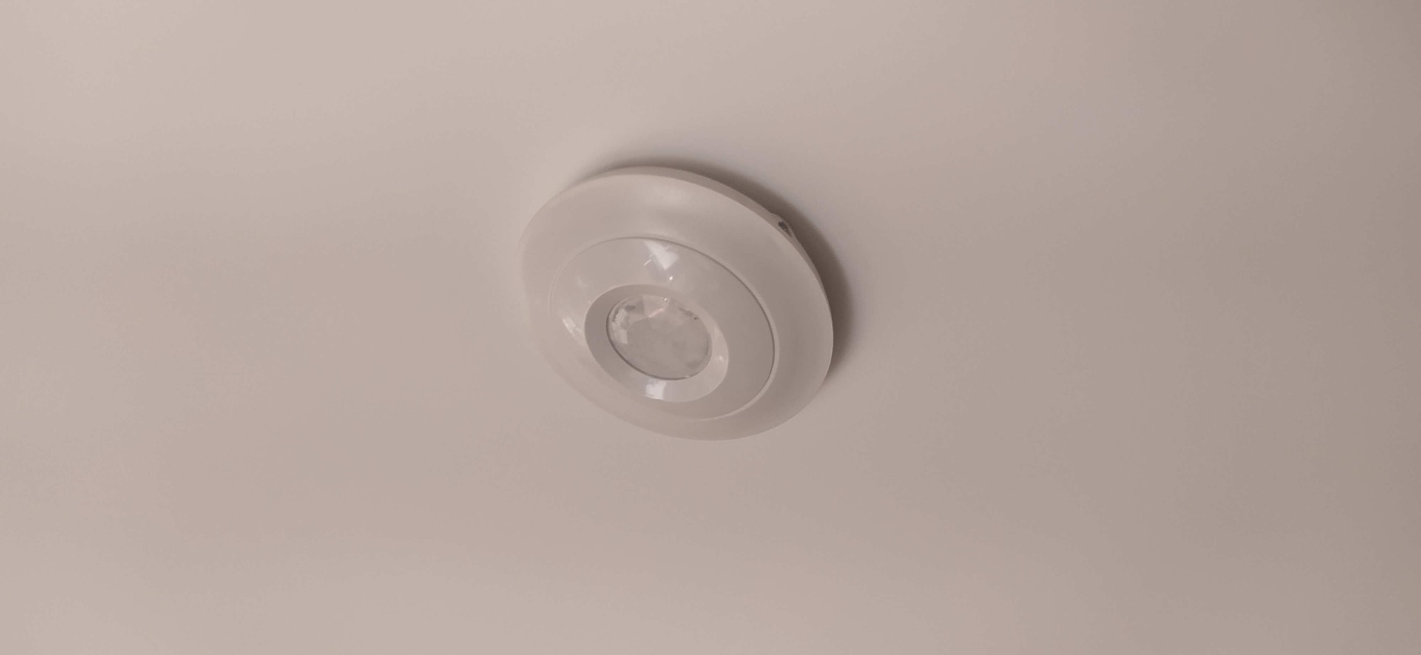 Zamontowany, biały czujnik ruchu na suficie. Okrągły kształt, widoczne elementy detekcji. Minimalistyczny design.