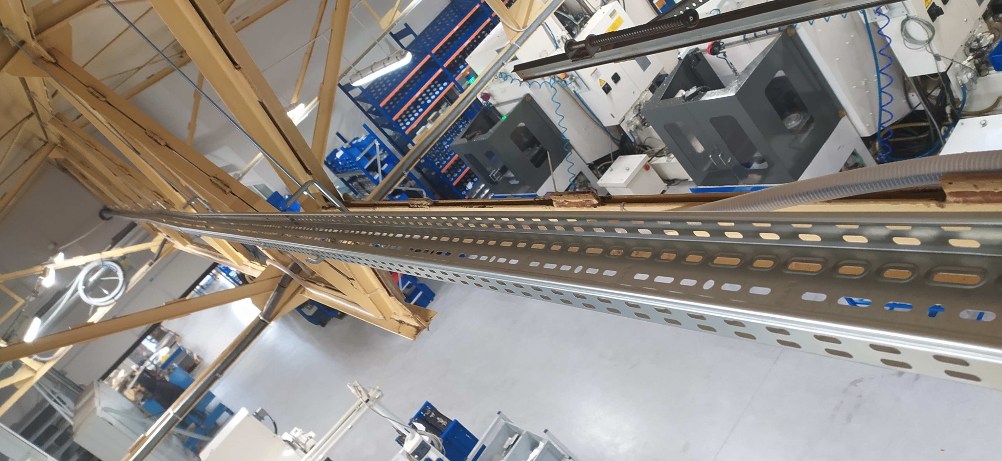 Wnętrze hali produkcyjnej z metalowymi regałami, widok z góry. W tle maszyny i niebieskie półki regałowe. Konstrukcja drewniana pod sufitem. Zdjęcie obrócone.