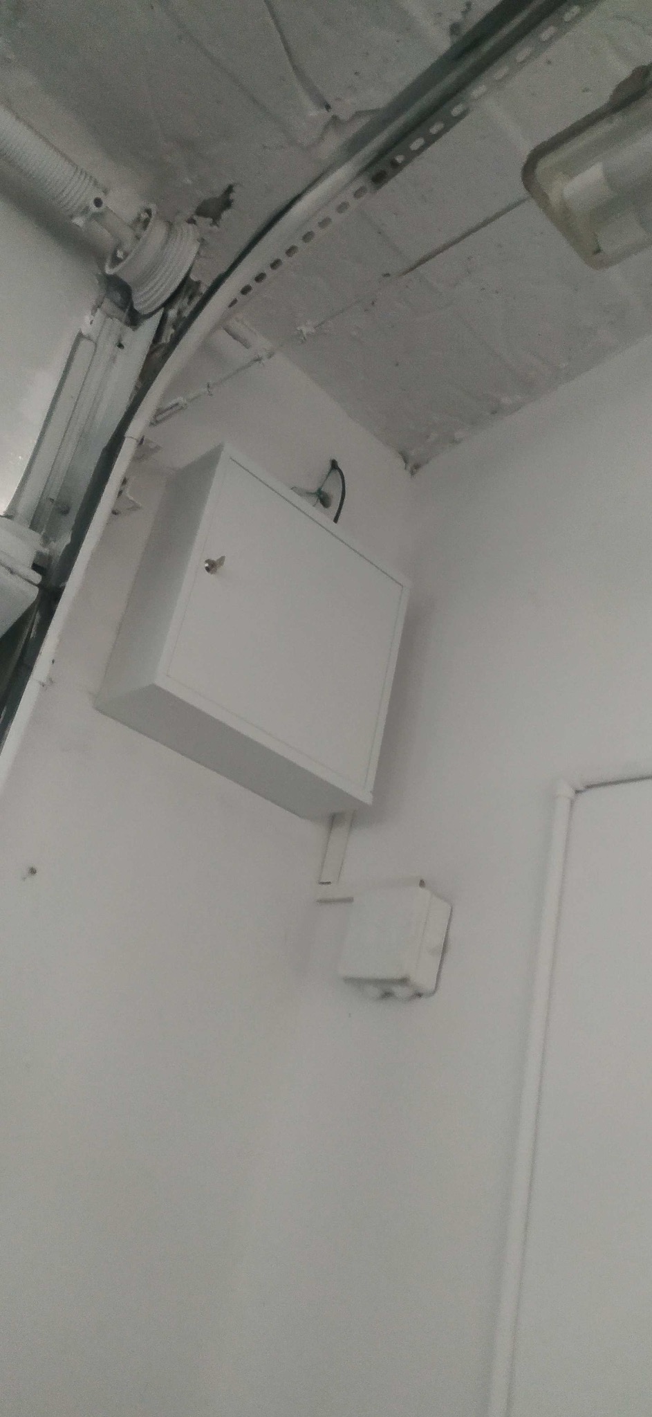 Biała skrzynka elektryczna z kablem na ścianie, widoczne rury instalacyjne pod sufitem. Całość w jasnej tonacji, ujęcie z dołu.