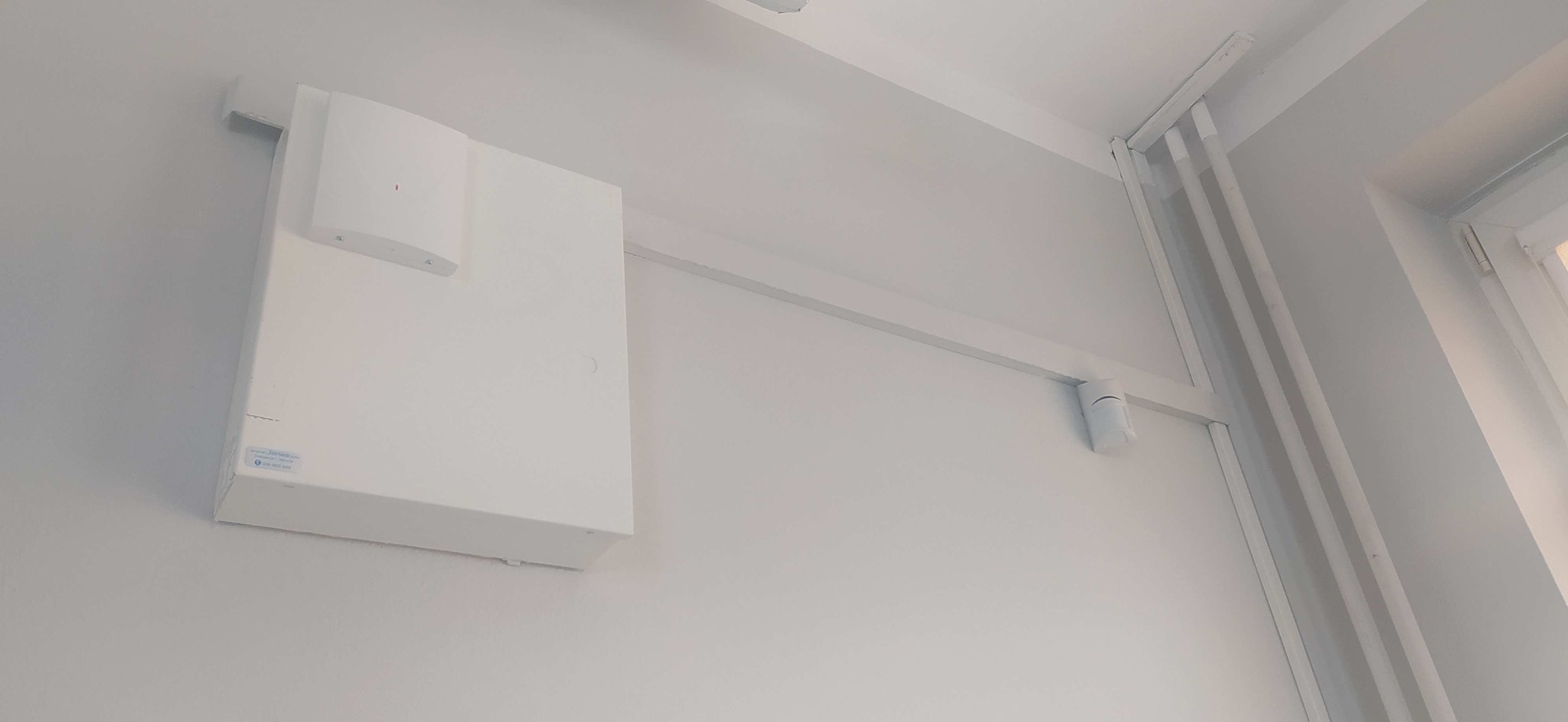 Biała centrala alarmowa z czujnikiem ruchu na ścianie, widoczne rury przy oknie. Minimalistyczny system zabezpieczeń w jasnym wnętrzu.