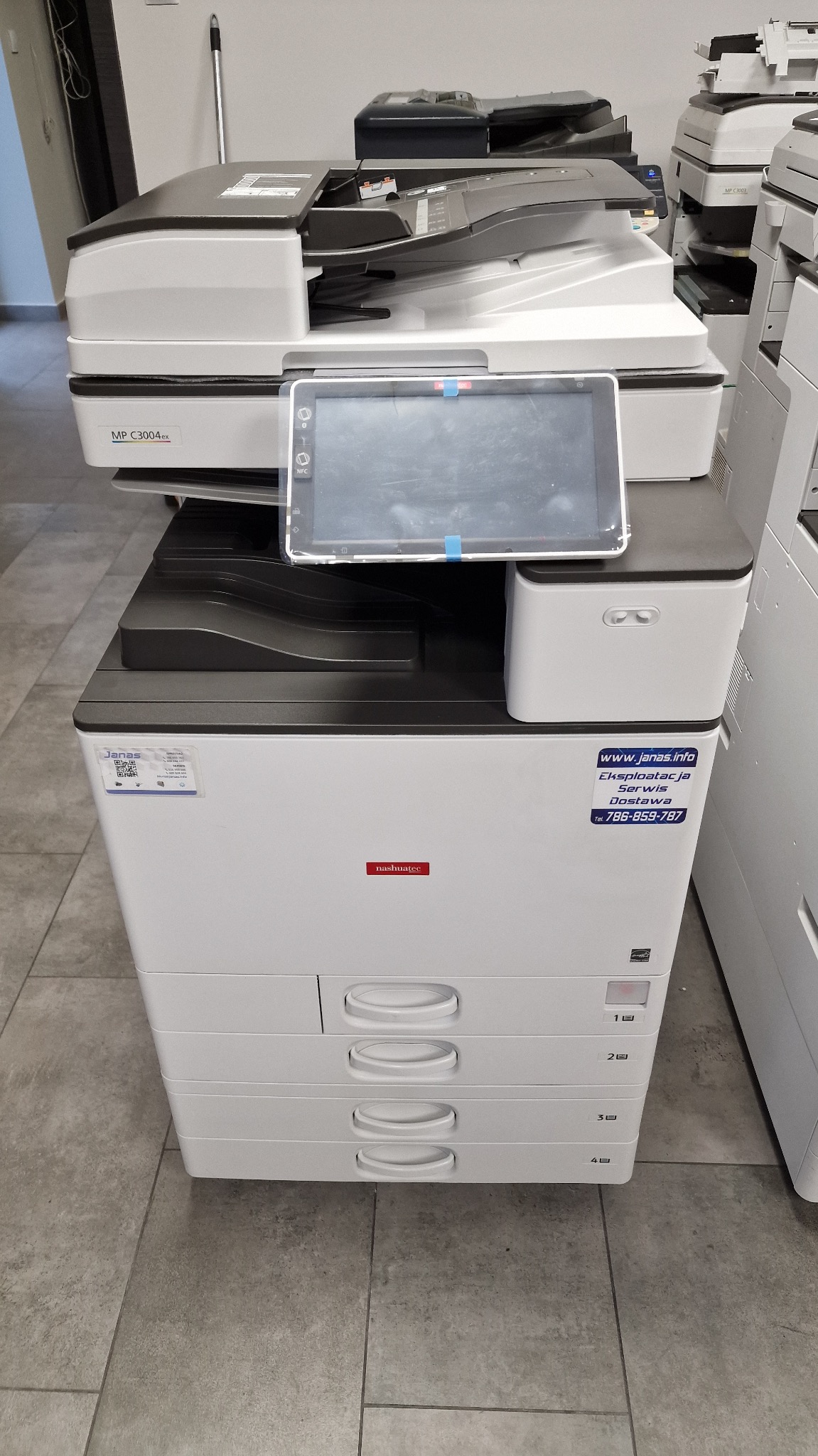 Biała kserokopiarka Nashuatec z czterema szufladami i panelem dotykowym, widoczna naklejka z danymi kontaktowymi serwisu. W tle inne urządzenia.