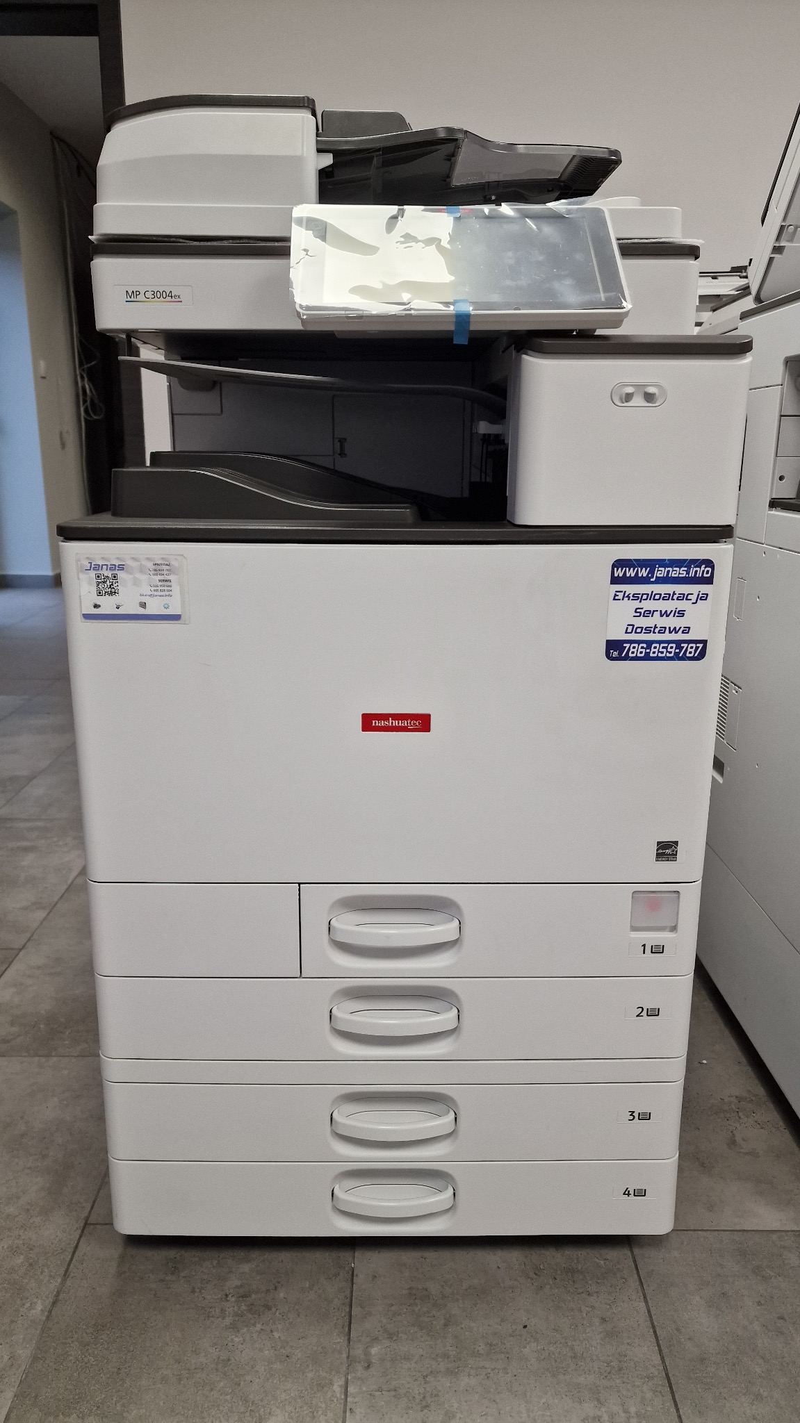 Wielofunkcyjna kserokopiarka Nashuatec MP C3004ex z naklejkami informacyjnymi firmy Janas, widok z przodu na szarej podłodze.
