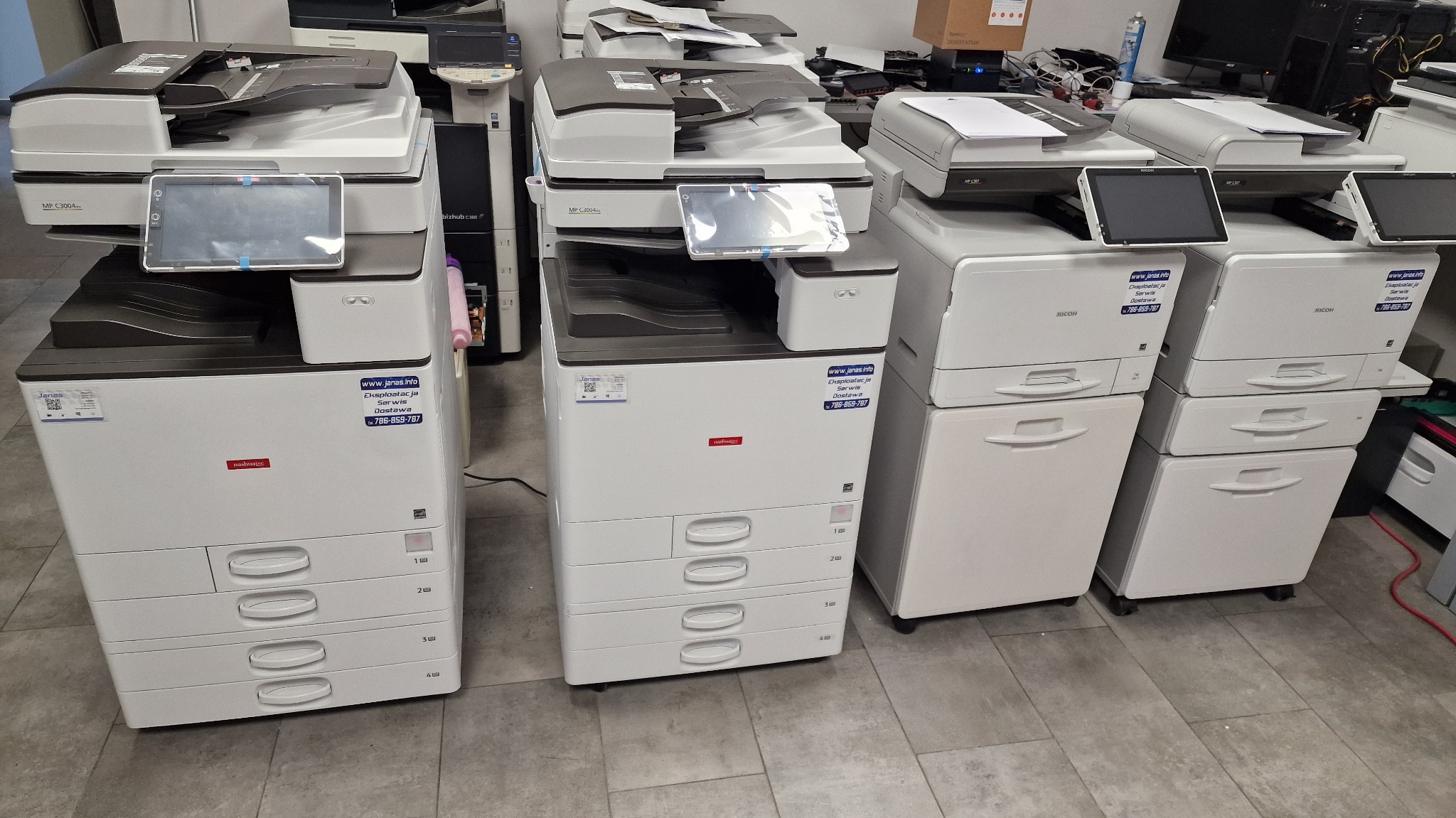 Rząd używanych, wielofunkcyjnych kserokopiarek A4, różnych modeli i marek, z widocznymi panelami dotykowymi, w pomieszczeniu biurowym przed serwisem.