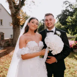 Para młoda w plenerze: panna młoda w sukni z kwiatowymi aplikacjami i welonem, pan młody w czarnym smokingu z bukietem białych kwiatów, w tle budynek i zieleń.