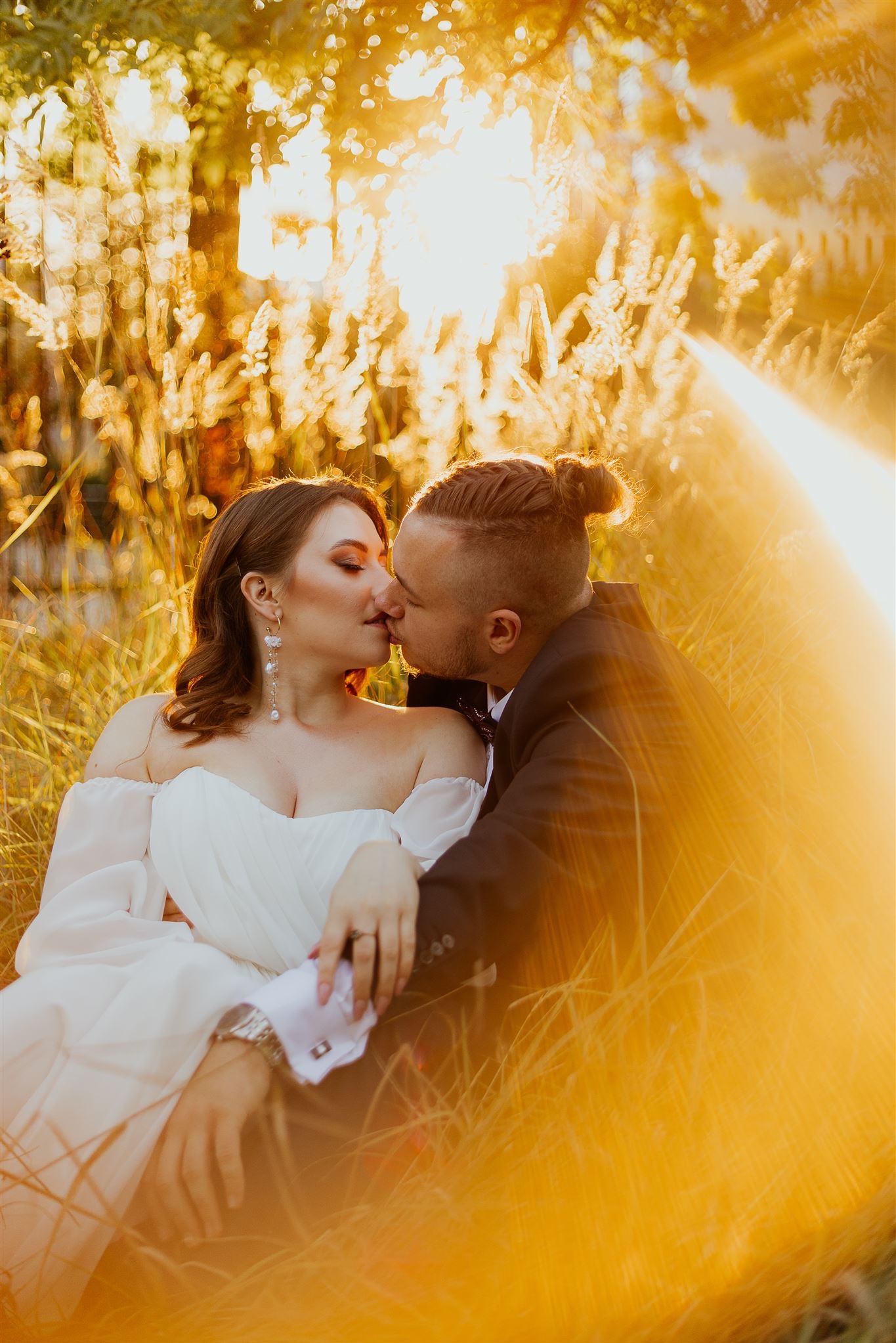 Para młoda w objęciach, pocałunek na tle złocistej trawy w promieniach zachodzącego słońca, panna młoda w sukni z odkrytymi ramionami i wiszącymi kolczykami, pan młody w czarnym garniturze z muchą.