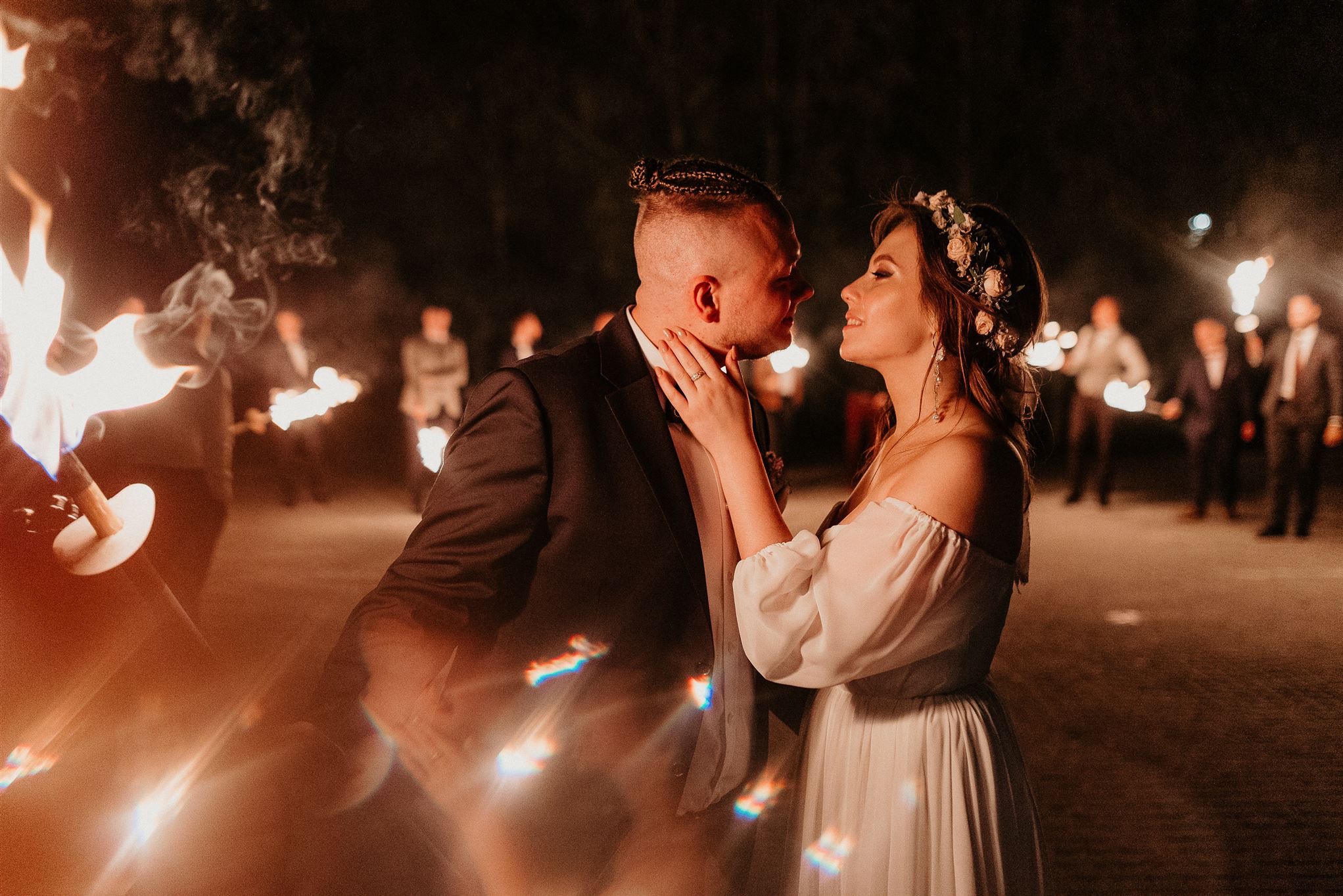 Para młoda wpatrzona w siebie, oświetlona blaskiem pochodni trzymanych przez gości weselnych w tle, panna młoda z wiankiem na głowie dotyka twarzy pana młodego.