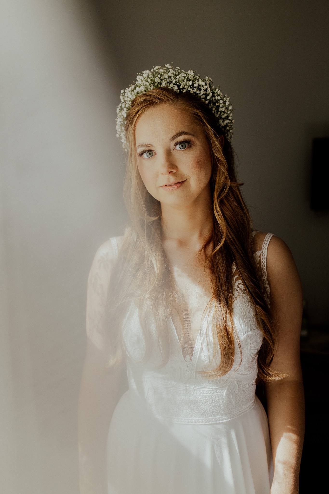 Portret panny młodej w białej sukni z koronkowymi detalami i wiankiem z gipsówki, oświetlonej miękkim światłem wpadającym przez okno.