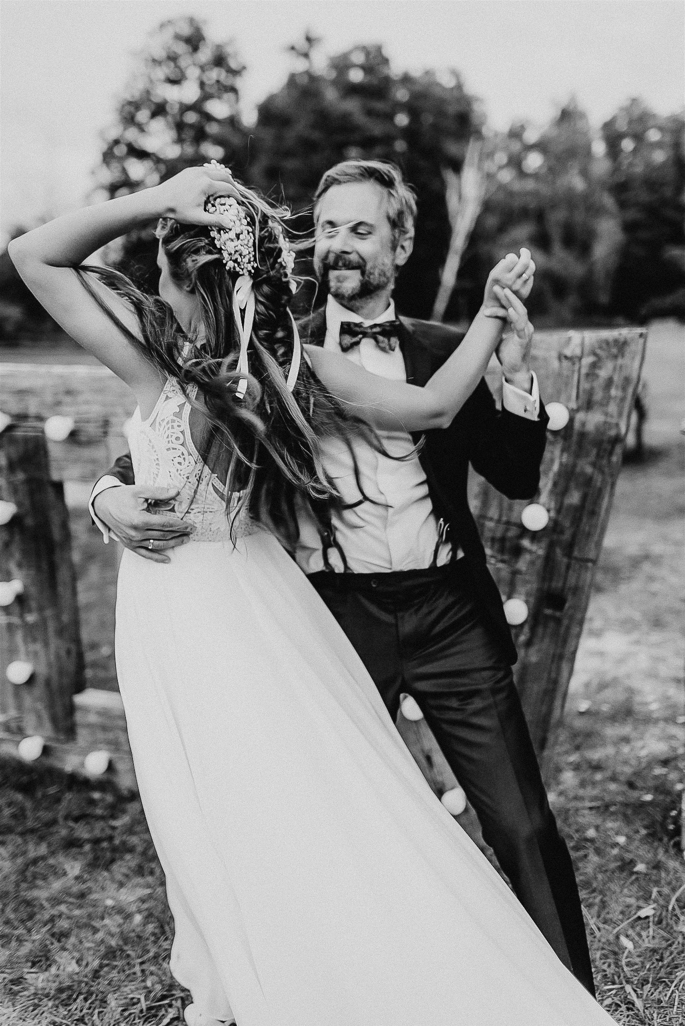 Czarno-białe zdjęcie pary młodej tańczącej na zewnątrz; panna młoda w koronie z kwiatów i sukni z koronkowymi detalami na plecach, pan młody w muszce i szelkach, oświetlona dekoracja za nimi.