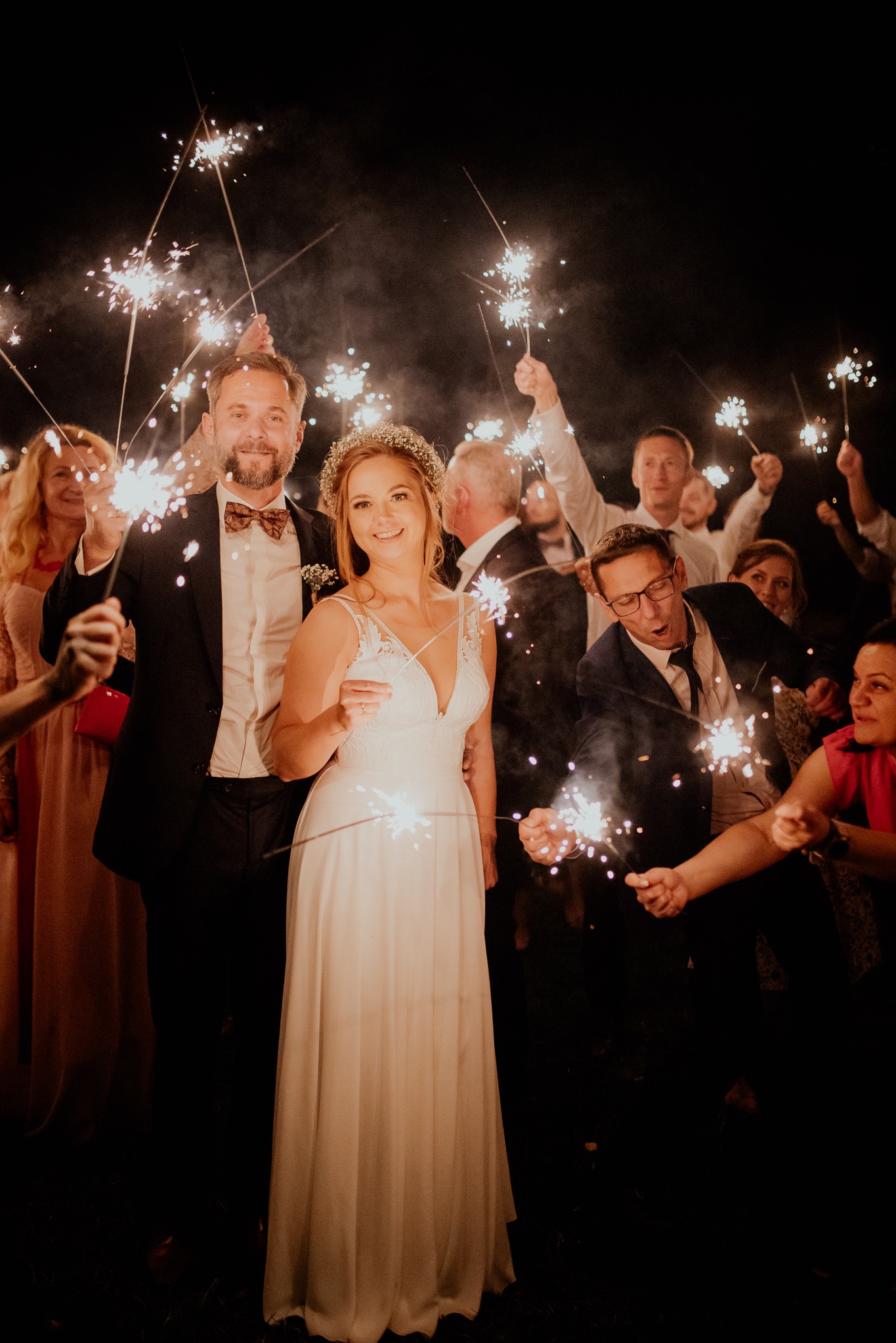 Para młoda w otoczeniu gości weselnych trzymających zapalone zimne ognie w ciemności, mężczyzna w muszce, kobieta w wianku i sukni ślubnej.